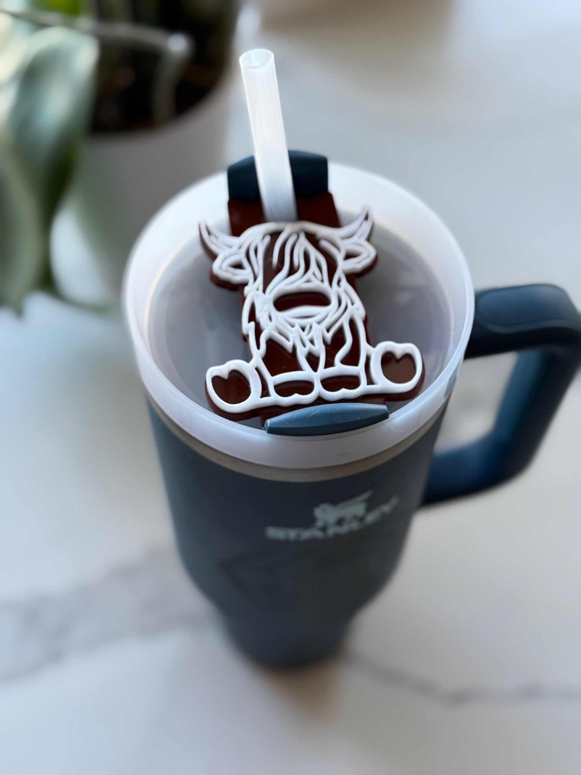 A dark gray Stanley tumbler filled with iced coffee sits on a white surface. The cup features a whimsical cow-shaped lid attachment with a straw through it. A blurred plant is in the background, adding a touch of greenery.