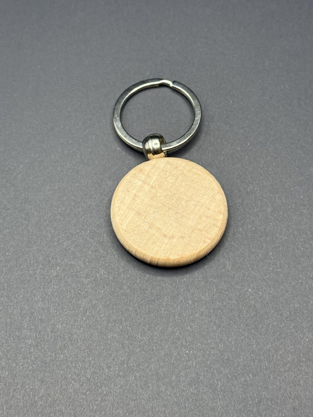 A round, wood-textured keychain with a silver metal ring attached. The keychain is flat and smooth, resting on a plain dark gray surface.