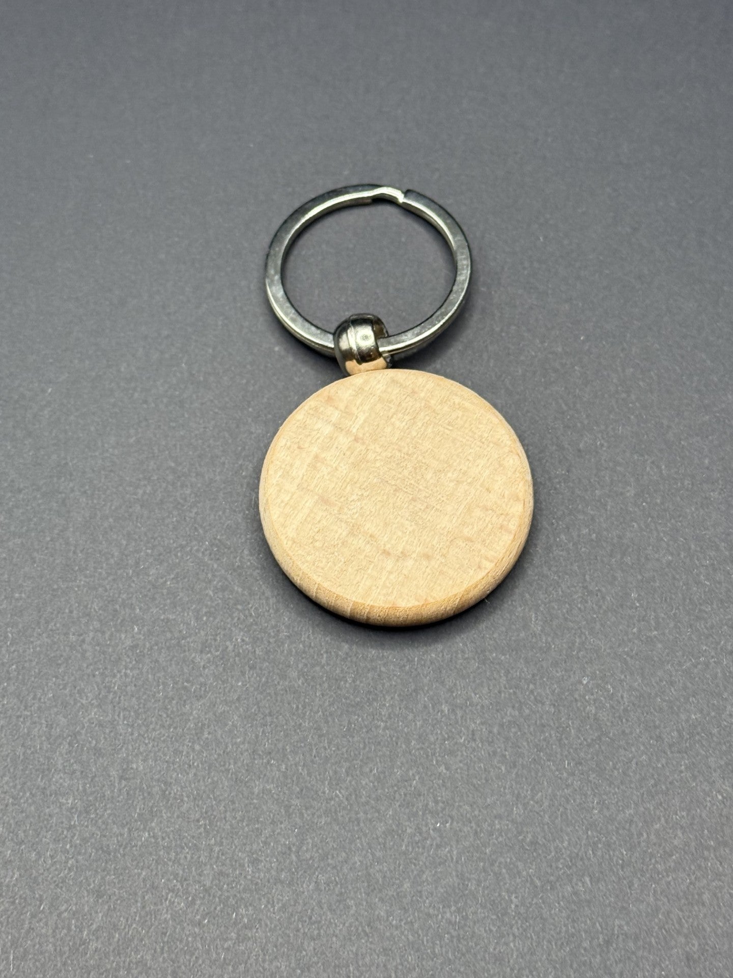 A round, wood-textured keychain with a silver metal ring attached. The keychain is flat and smooth, resting on a plain dark gray surface.