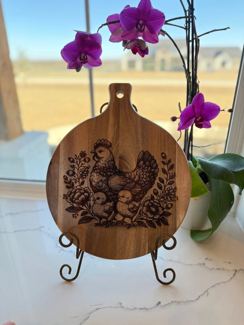 The Patriotic 3D Eagle Flag engraved Acacia Wood Cutting Board features intricate details and sits elegantly on a stand. Placed on a white marble surface near a window with purple orchids, it creates a striking, rustic scene enhanced by the natural beauty of acacia wood.