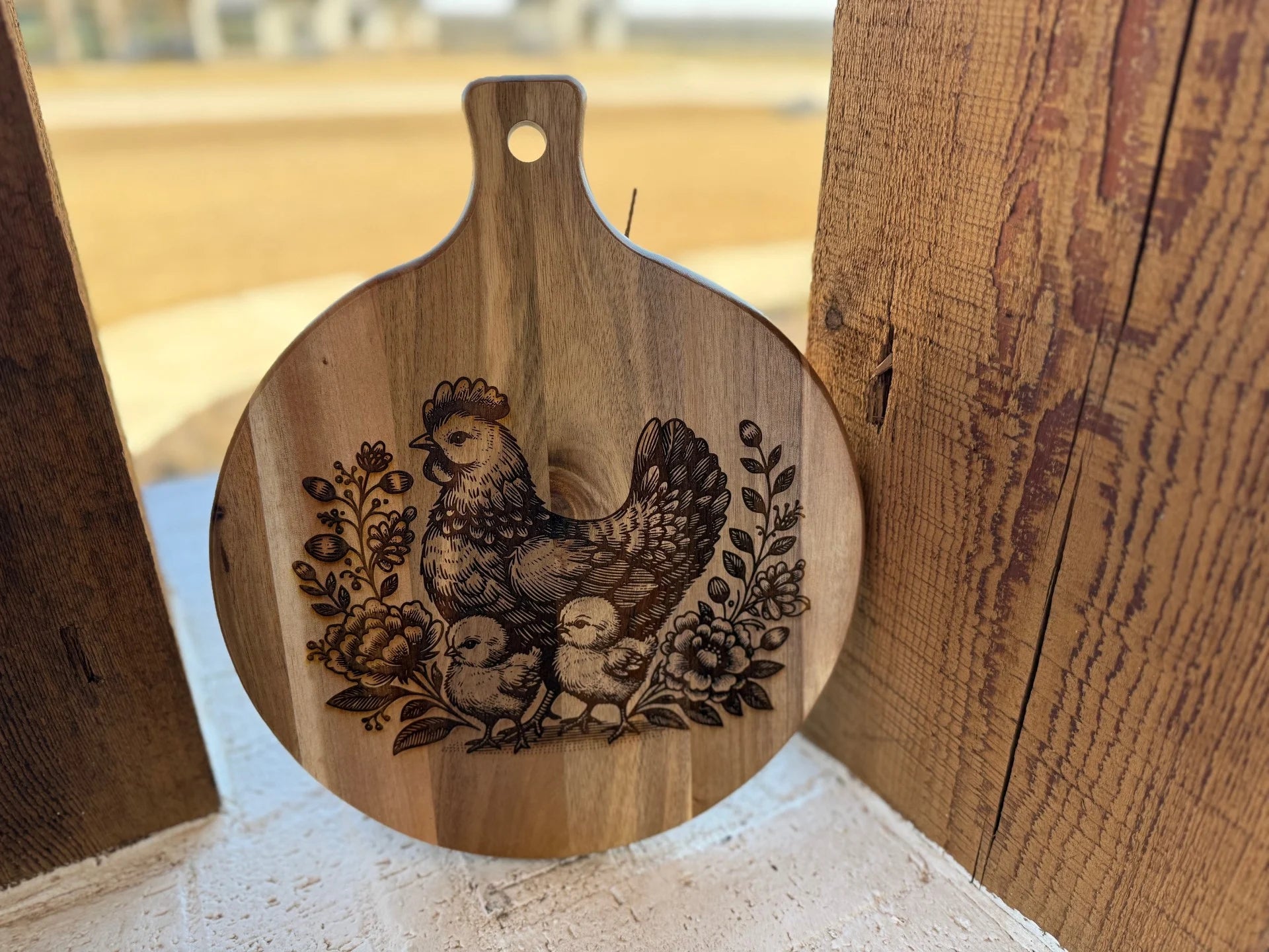 An acacia wood cutting board features a patriotic 3D eagle flag design. It rests on a rustic wooden surface, with an outdoor setting highlighted by blurred grass and sky in the background.