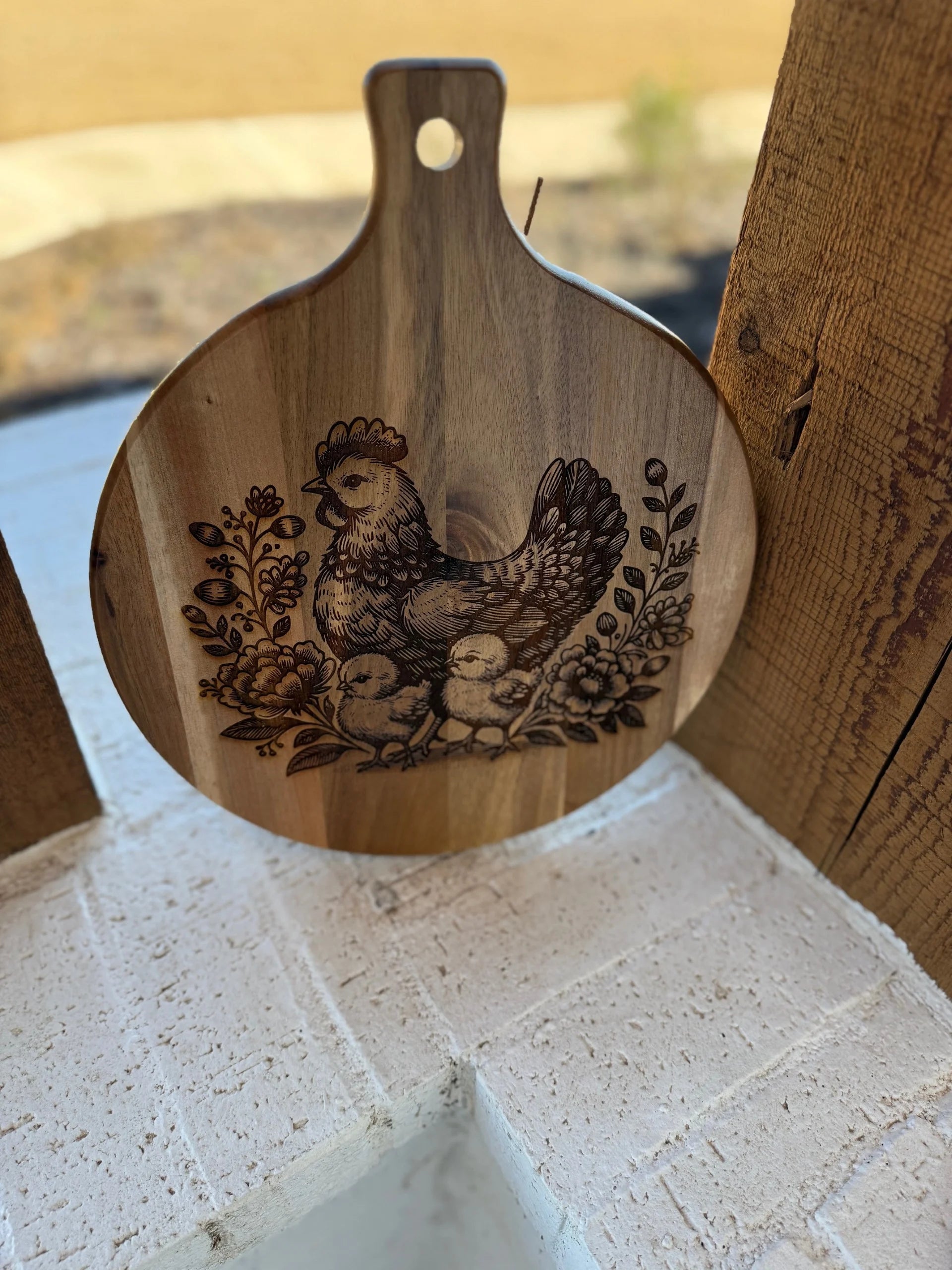 The Patriotic 3D Eagle Flag engraved Acacia Wood Cutting Board hangs on a wooden wall, showcasing an eagle design. The outdoor view includes grass and a white brick ledge, and the round board features a handle with a hanging hole.