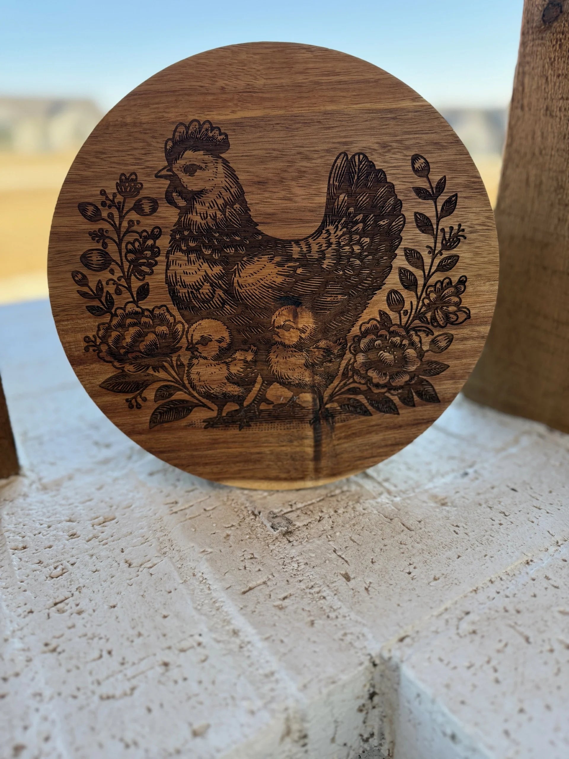 An acacia wood cutting board engraved with a detailed scene of a mother hen and chicks amid flowers, set against a blurred outdoor background and displayed on a white brick surface.