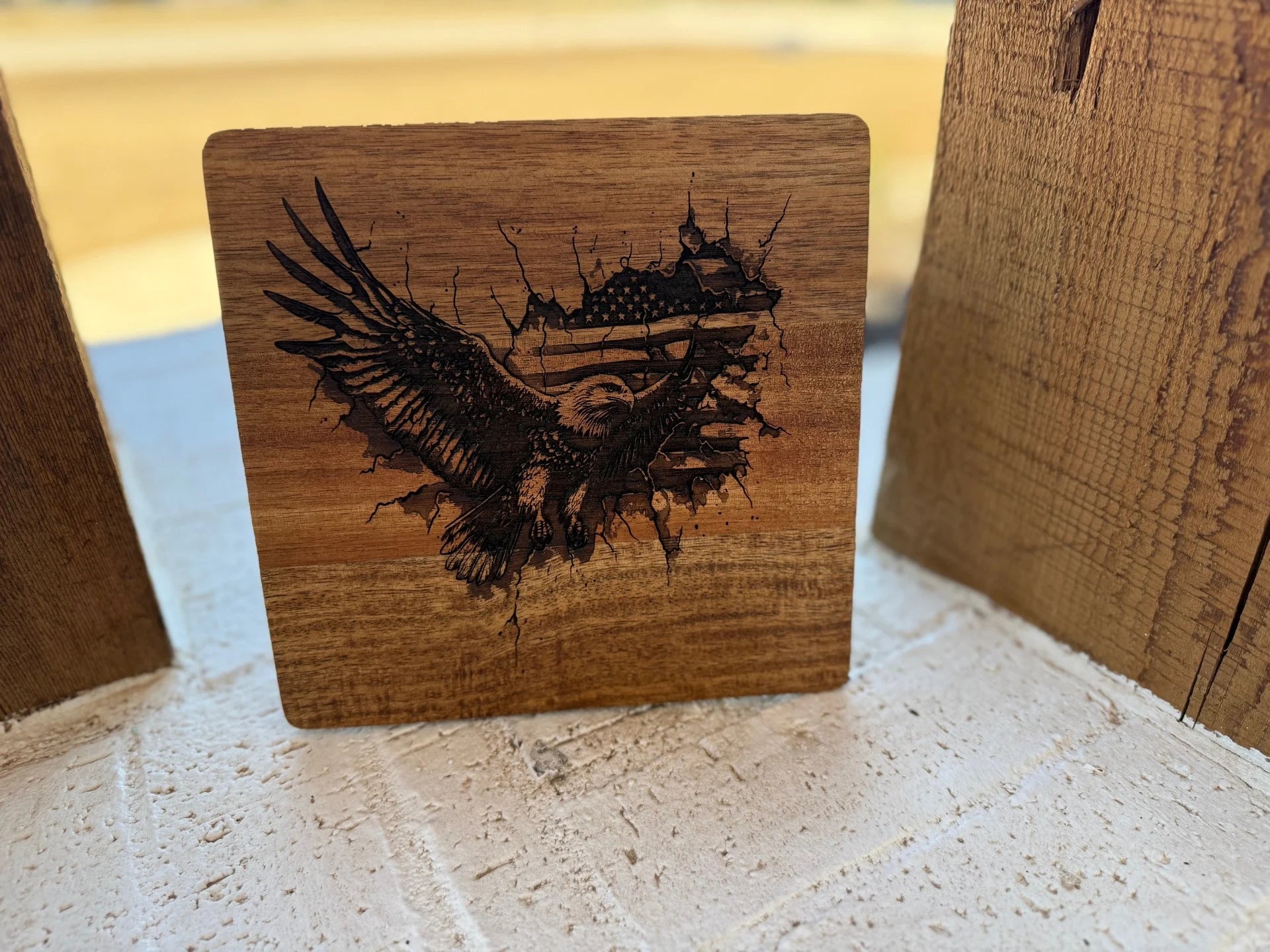 The Patriotic 3D Eagle Flag engraved Acacia Wood Cutting Board features an eagle in flight over an American flag backdrop. The cracked surface effect adds depth, while natural light accentuates the details against its textured white and wooden base.