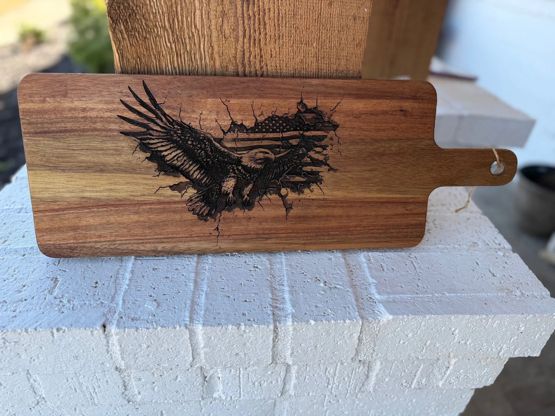 The Patriotic 3Eagle engraved Acacia Wood Cutting Board, featuring an eagle with outstretched wings and an American flag design, combines flair with functionality. Displayed on a white brick ledge with a rustic wooden background, it has a handle and a small hole for easy hanging.