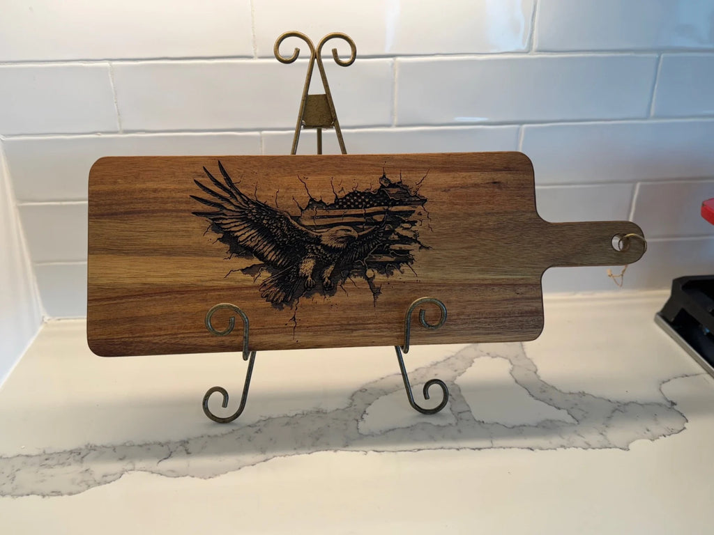 The Patriotic Eagle-engraved Acacia Wood Cutting Board beautifully rests on a decorative metal stand against a marbled white countertop and white tiled backsplash, evoking patriotic pride with its eagle design.