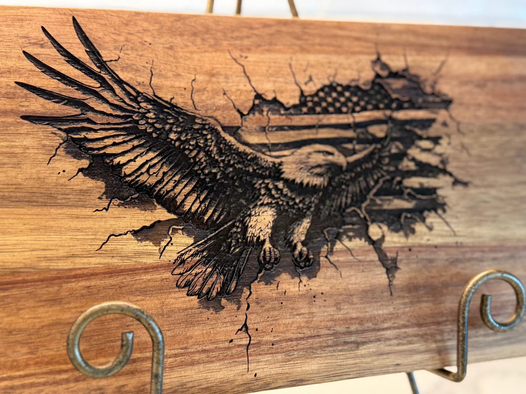 The Patriotic 3Eagle engraved Acacia Wood Cutting Board features a bald eagle in flight with wings spread wide against an integrated American flag, creating a three-dimensional effect. Displayed on a metal stand, this sturdy piece resembles an elegant cutting board.