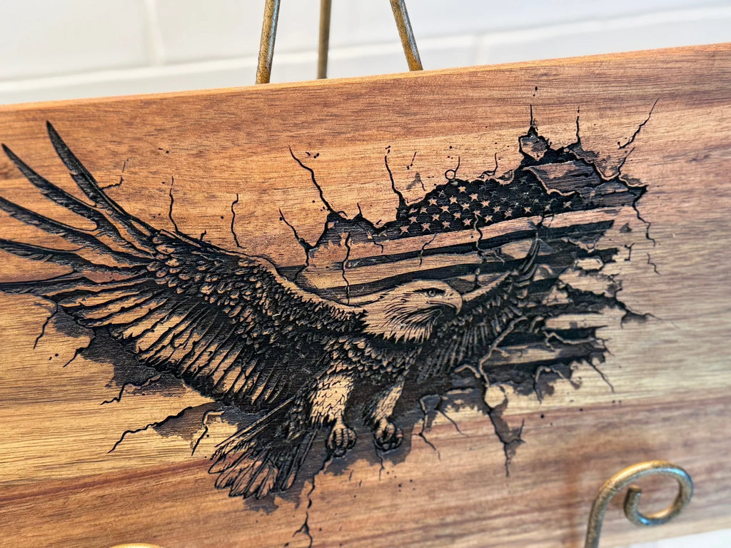 The Patriotic 3Eagle Engraved Acacia Wood Cutting Board showcases an eagle with outstretched wings against the American flag, with its natural grain enhancing the majestic design, elegantly set on a metal stand and shown against a white brick background.
