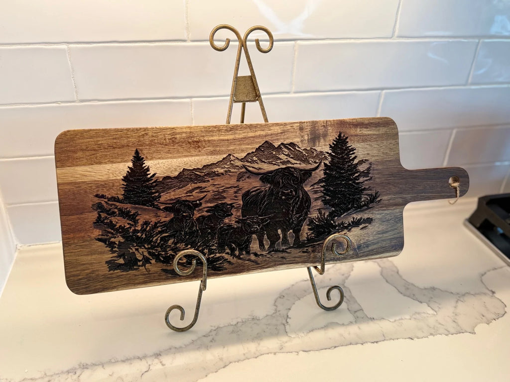 BBQ Grill Chart engraved Acacia Wood Cutting Board displayed on a decorative metal stand, set against a white tiled wall and marble countertop, featuring an intricate mountain, tree, and bison with calf scene.
