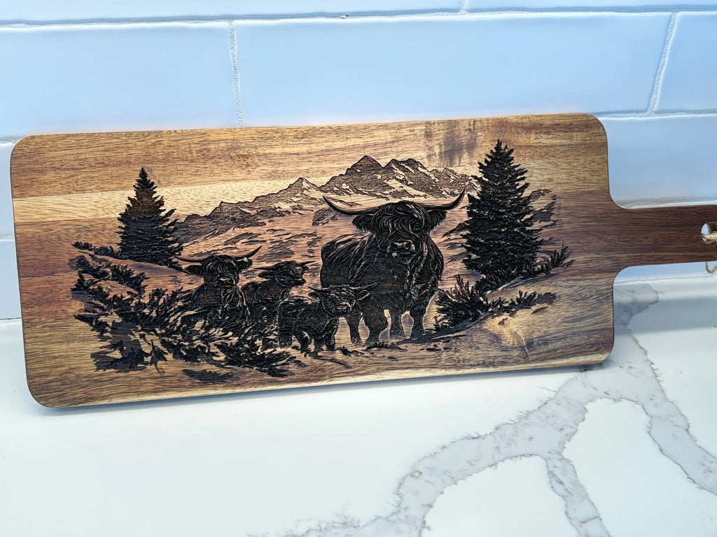 The BBQ Grill Chart engraved Acacia Wood Cutting Board features a highland cow and two calves among trees and mountain silhouettes. The light brown acacia wood contrasts with the dark engraving, displayed on a white marbled surface against a tiled wall.