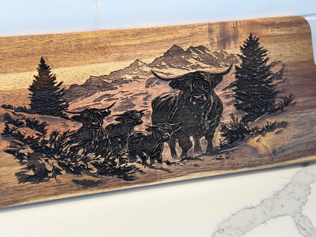 BBQ Grill Chart engraved Acacia Wood Cutting Board features a scenic landscape with mountains, trees, a highland cow, and three calves among grass. It sits elegantly on a white marble countertop, blending natural artistry and timeless kitchen charm.