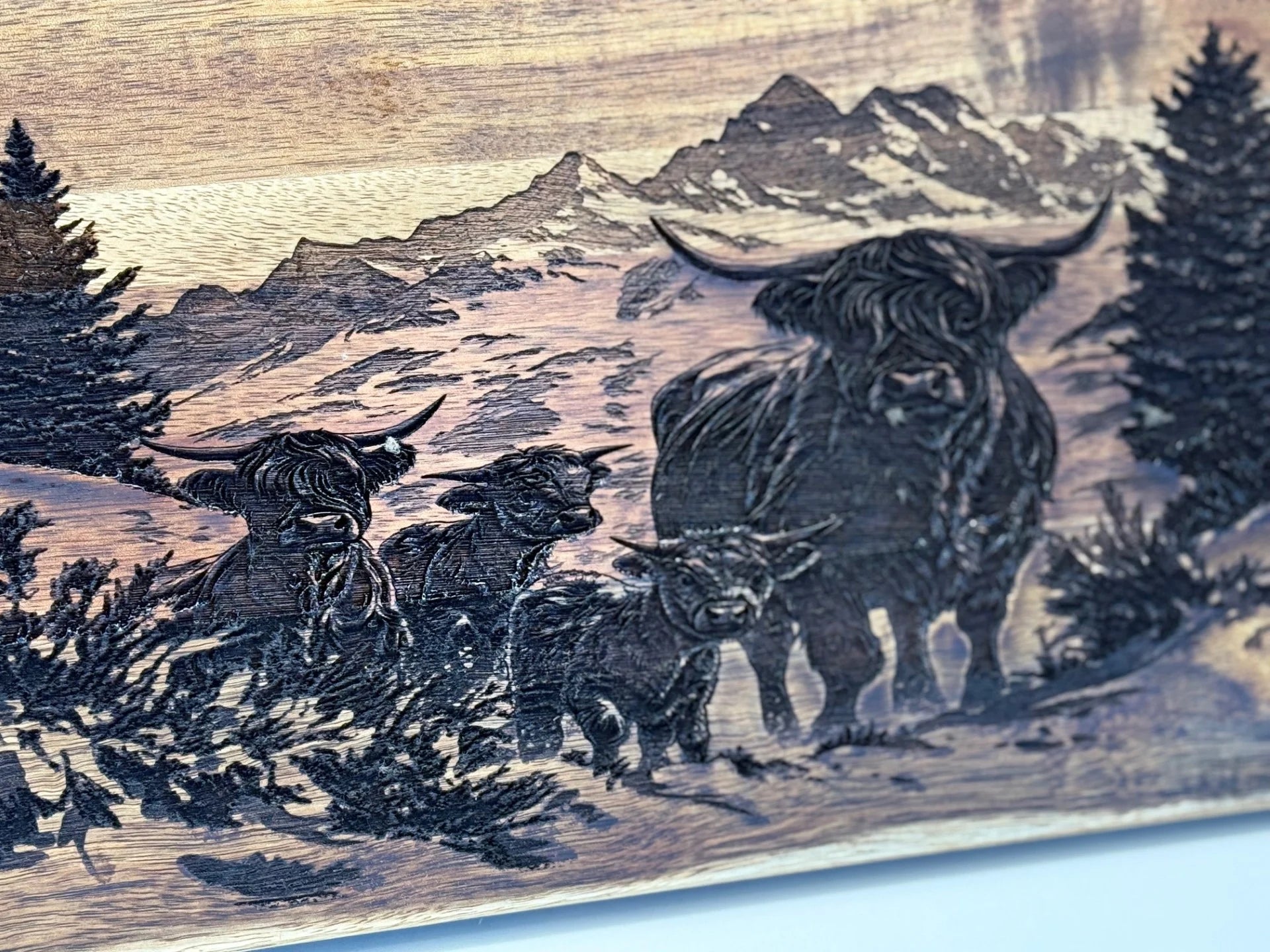 Engraved on the BBQ Grill Chart Acacia Wood Cutting Board, a Highland cow family—one large cow and three calves—stands against mountains. Intricate carving highlights their features amid trees and bushes, with peaks in the background.