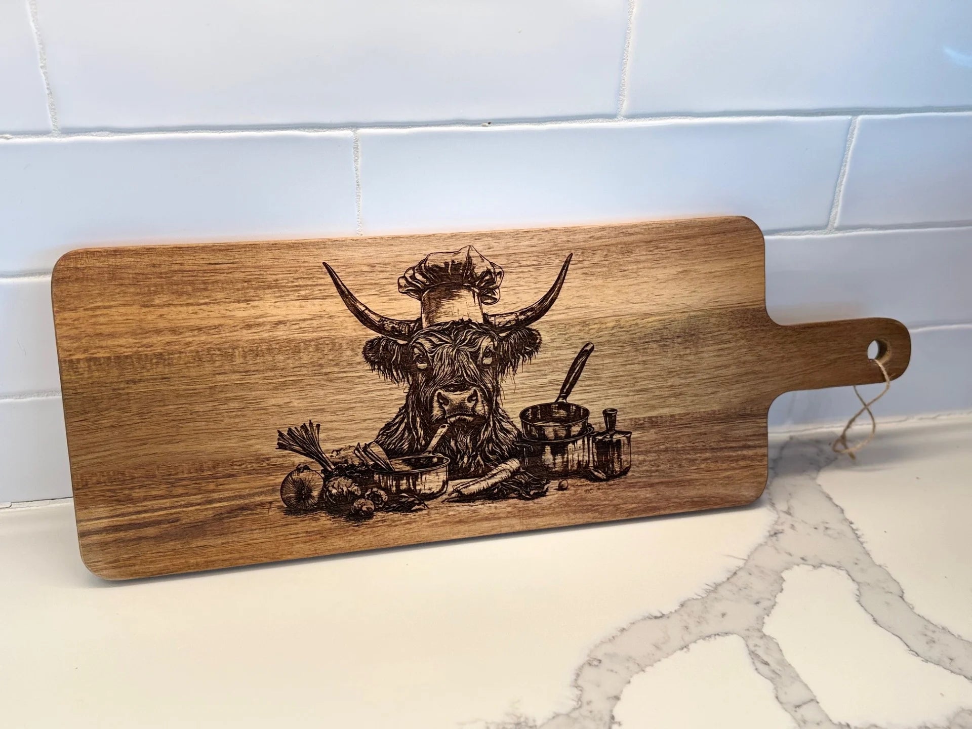 The BBQ Grill Chart engraved Acacia Wood Cutting Board showcases a cow wearing a chefs hat, with kitchen items like a bowl, vegetables, pepper grinder, and spoon. Set on a marble countertop with a white tiled backsplash, its ideal for preparing BBQ grill favorites.