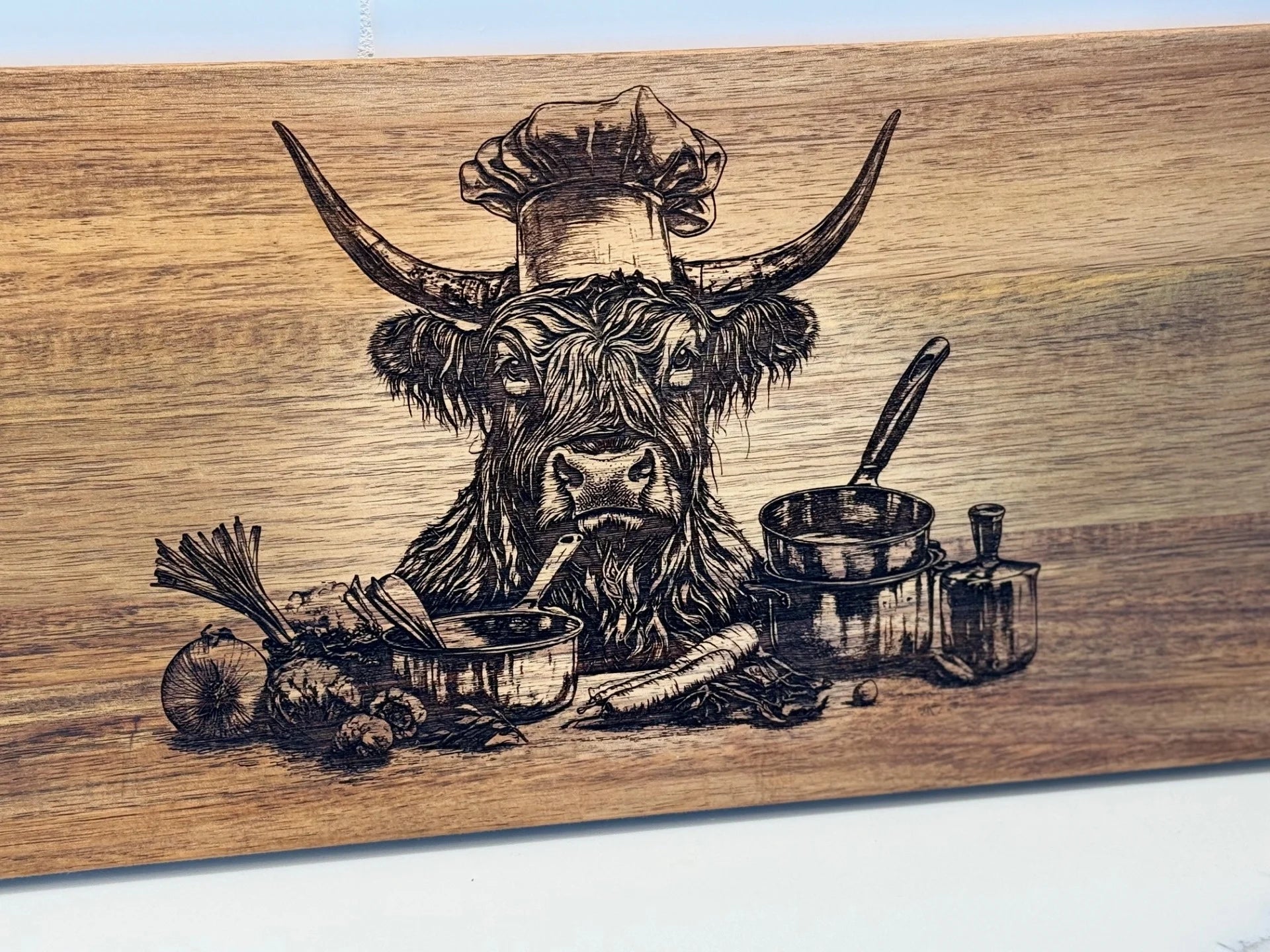 The BBQ Grill Chart engraved Acacia Wood Cutting Board features a Highland cow in a chefs hat, tasting from a spoon amidst vegetables and cooking pots, creating a humorous culinary scene with onions, carrots, and leafy greens.