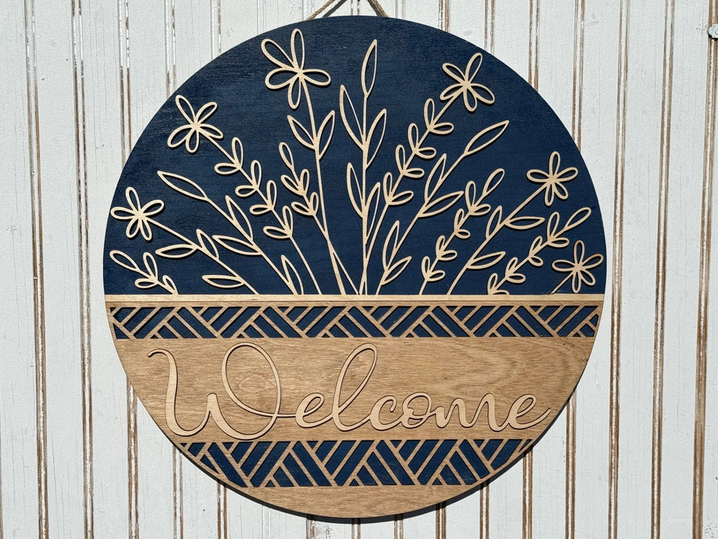 This round wooden Welcome Door Sign, Welcome Wreath, or Welcome Door Hanger boasts Welcome in elegant script. It features a dark blue top with floral cutouts and a light wood bottom with a geometric design, making it perfect against a striped textured wall.