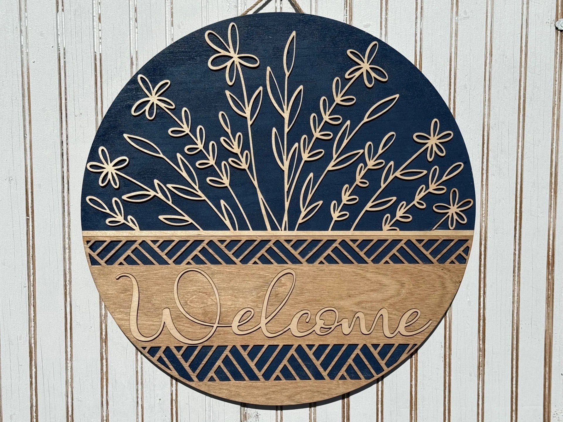 This round wooden Welcome Door Sign, Welcome Wreath, or Welcome Door Hanger boasts Welcome in elegant script. It features a dark blue top with floral cutouts and a light wood bottom with a geometric design, making it perfect against a striped textured wall.