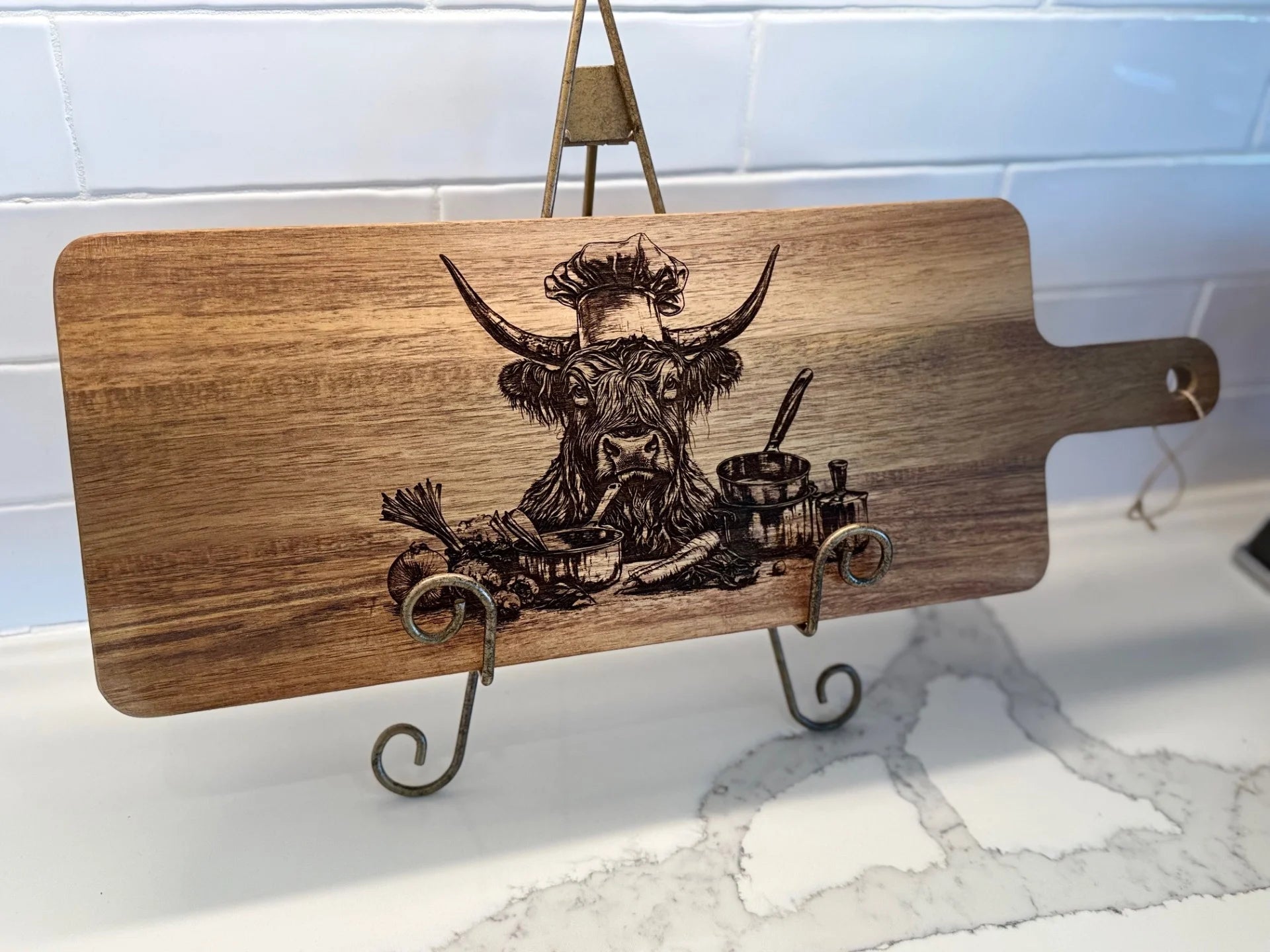 The BBQ Grill Chart engraved Acacia Wood Cutting Board has a bull in a chefs hat, cooking pots, a spoon, and herbs design. It includes a handle with a hole and sits on white marble against a tile backsplash, ideal for BBQ grill prep.