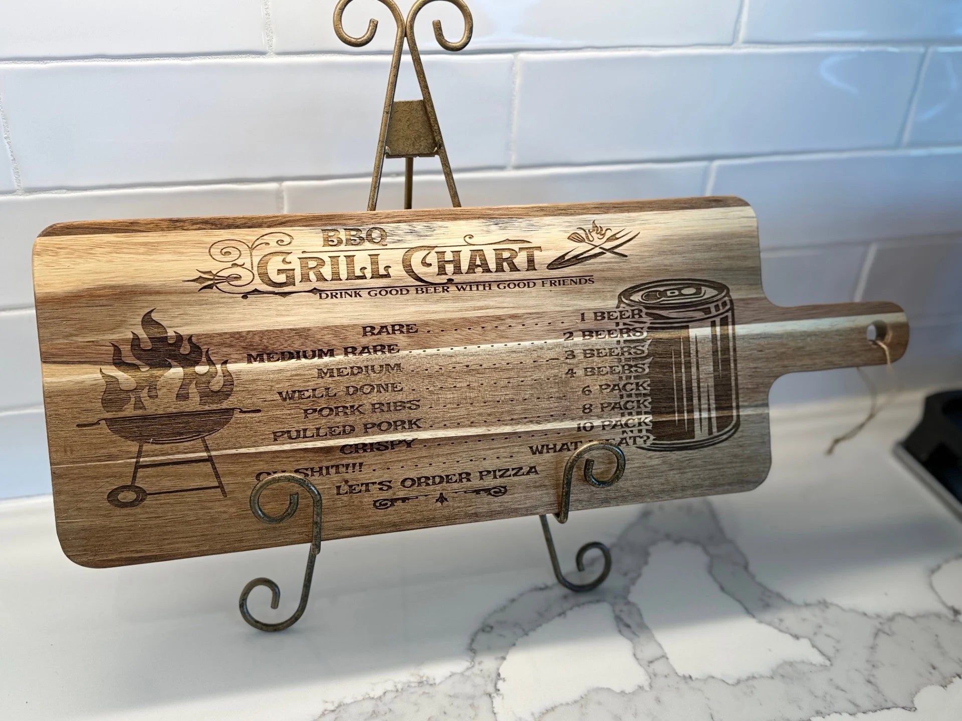 The BBQ Grill Chart engraved Acacia Wood Cutting Board features a metal stand, grill art with beer illustrations, and cooking levels from rare to pulled pork paired with beer suggestions. Text: “Drink good beer with good friends,” and “Shift: Let’s order pizza.” Set against a white tiled background.