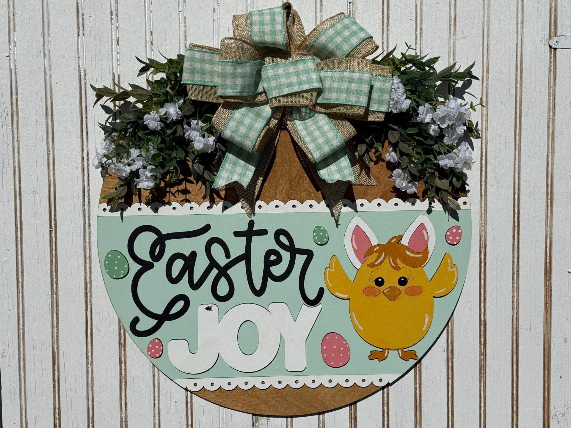 A festive Easter decoration on a white wooden background. It features the words Easter Joy with a cartoon yellow chick wearing bunny ears. Theres a large green gingham and burlap bow on top, surrounded by white flowers and greenery. Colorful Easter eggs dot the design.