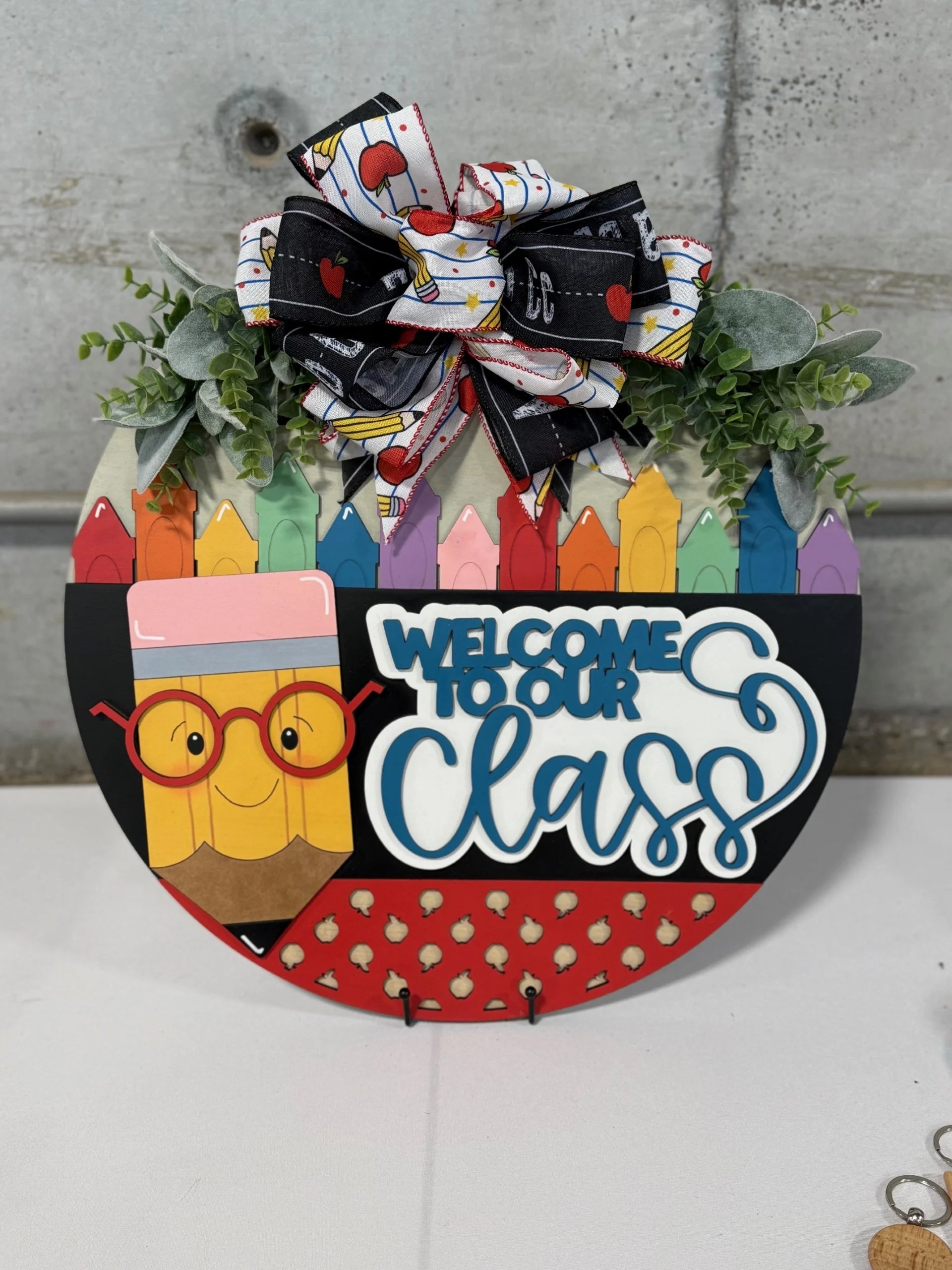 A decorative classroom sign shaped like a semi-circle features colorful cartoon pencils, a smiling pencil with glasses, the text Welcome to Our Class, and a bow with apple patterns. Greenery is tucked above the bow, set against a concrete background on a white surface.