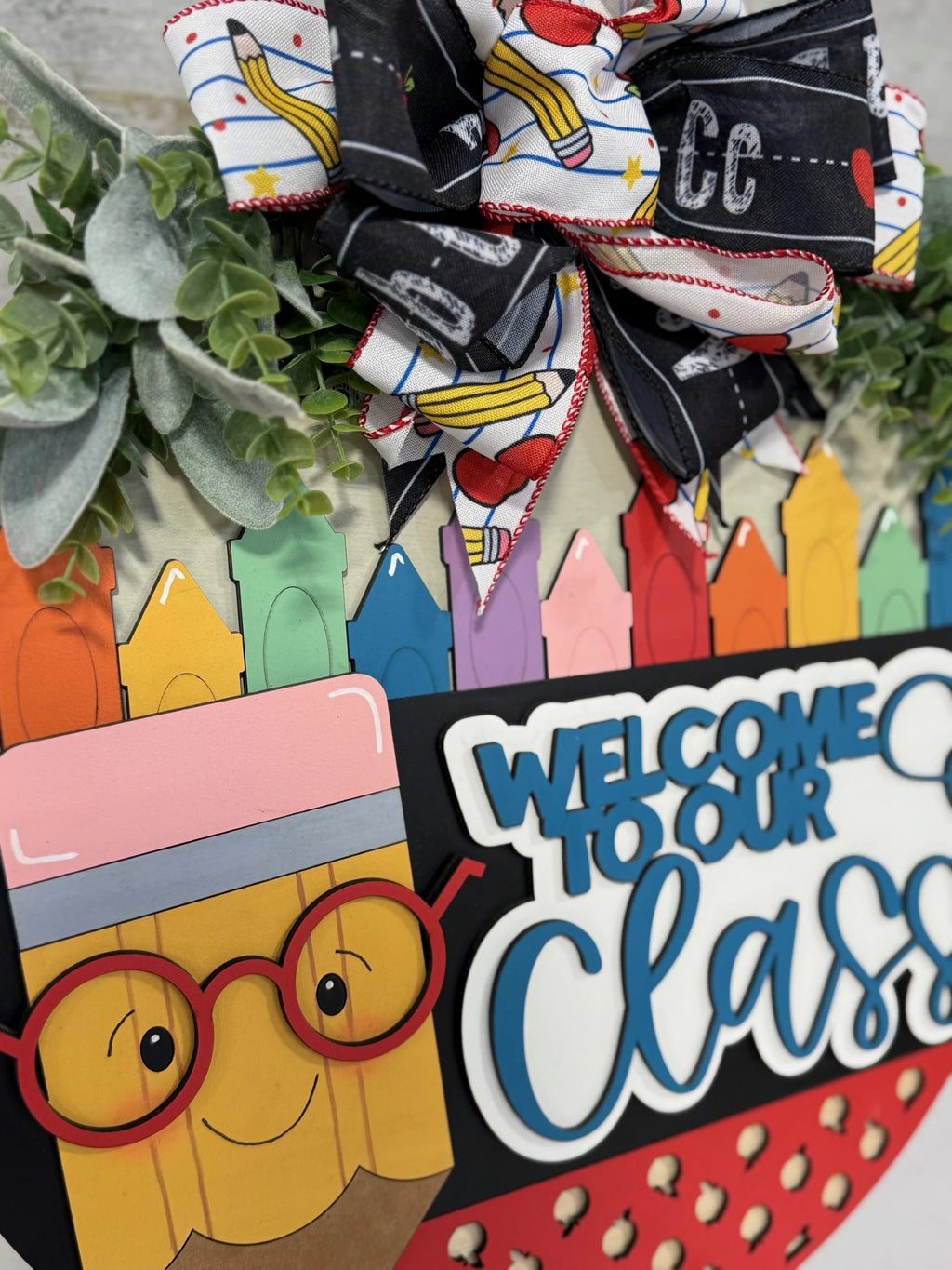 Close-up of a colorful classroom sign featuring a smiling pencil with red glasses. Above it is a Welcome to Our Class message in blue and white. The sign is decorated with a bow made of ribbons and greenery, with a backdrop of multicolored crayons.