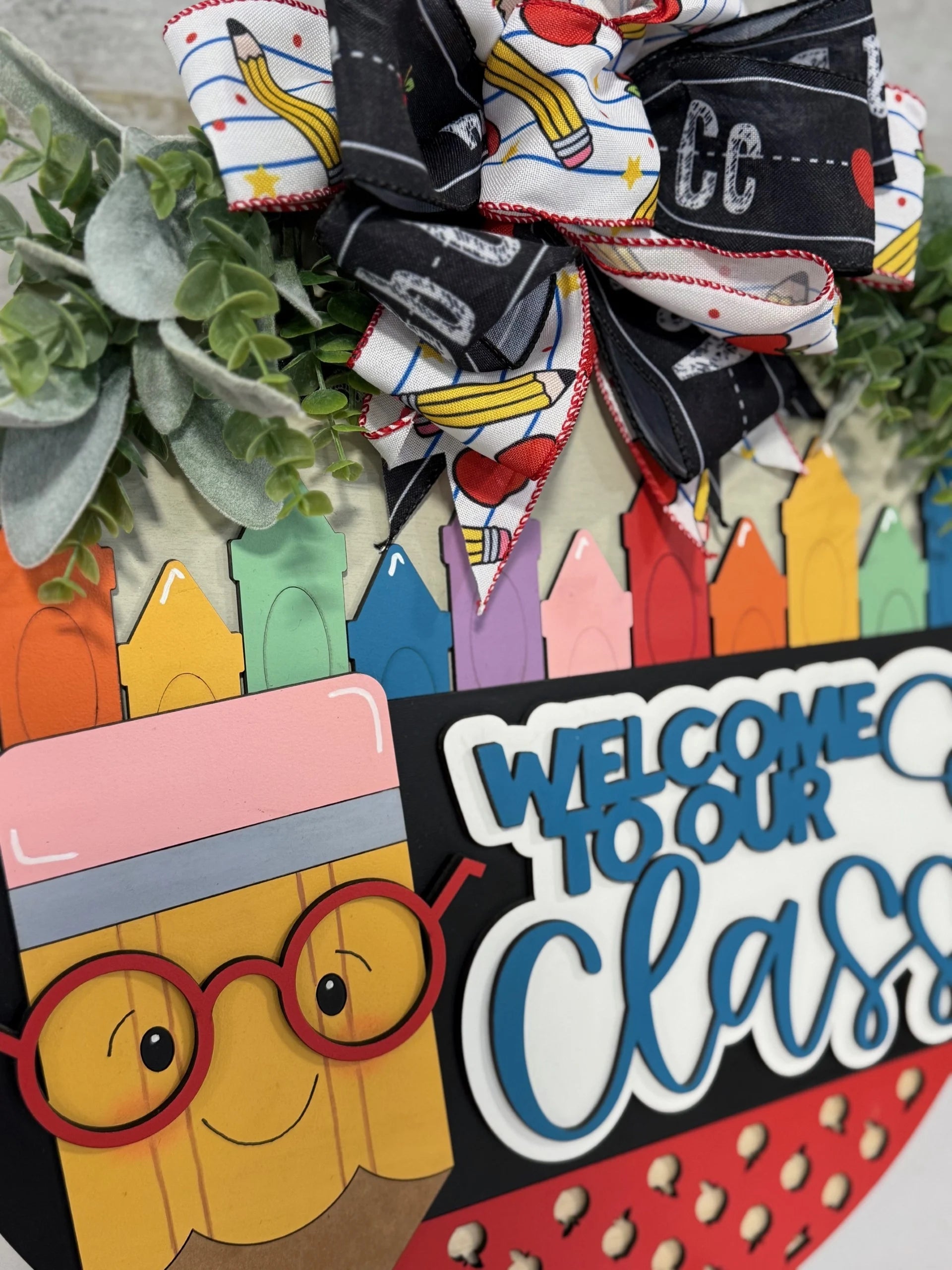 Close-up of a colorful classroom sign featuring a smiling pencil with red glasses. Above it is a Welcome to Our Class message in blue and white. The sign is decorated with a bow made of ribbons and greenery, with a backdrop of multicolored crayons.