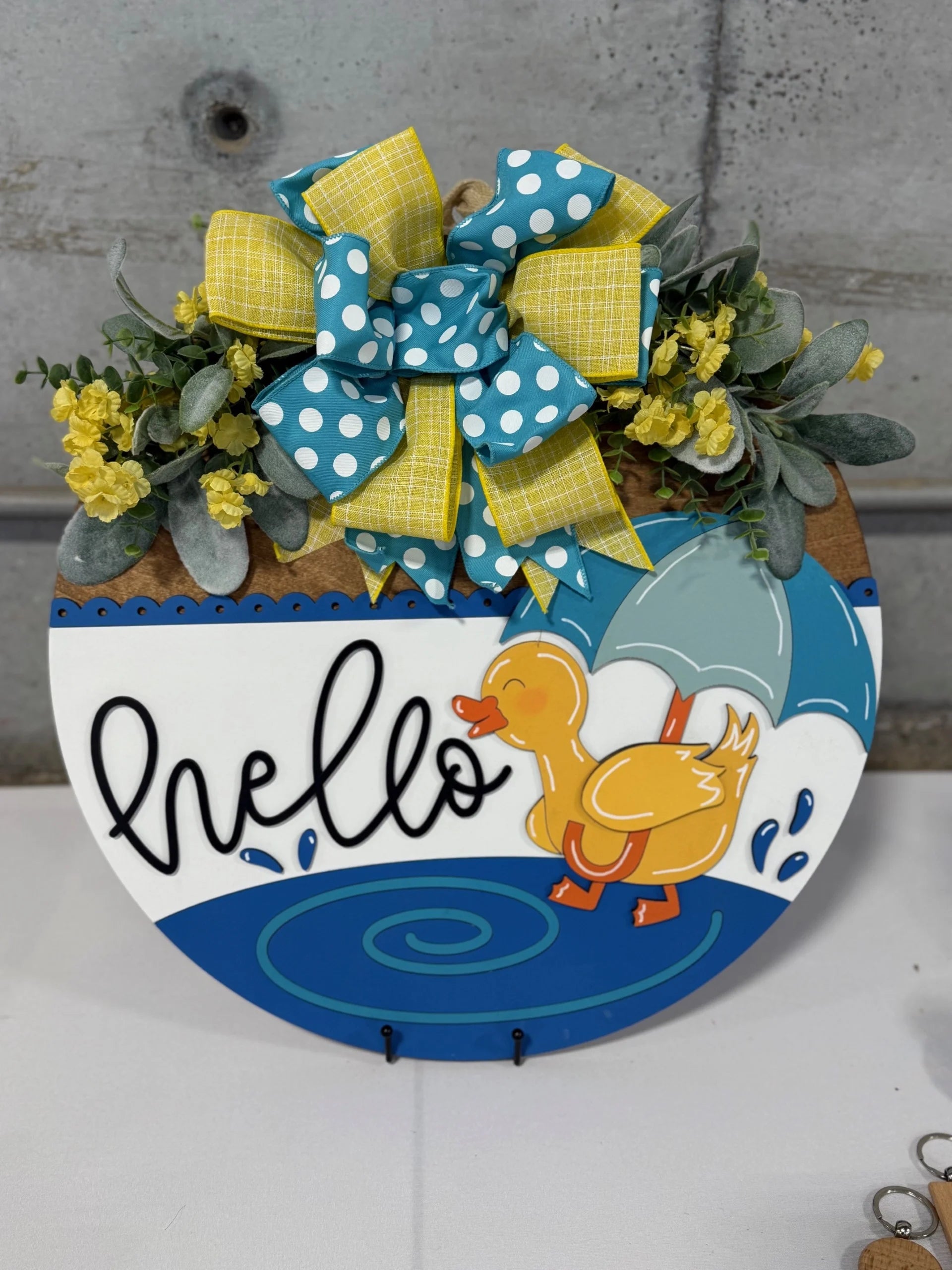 Round decorative sign with a cheerful yellow duck holding an umbrella. The word hello is written in black cursive. A large blue and yellow bow with polka dots and plaid patterns adorns the top, surrounded by green leaves and yellow flowers. Background is a concrete wall.