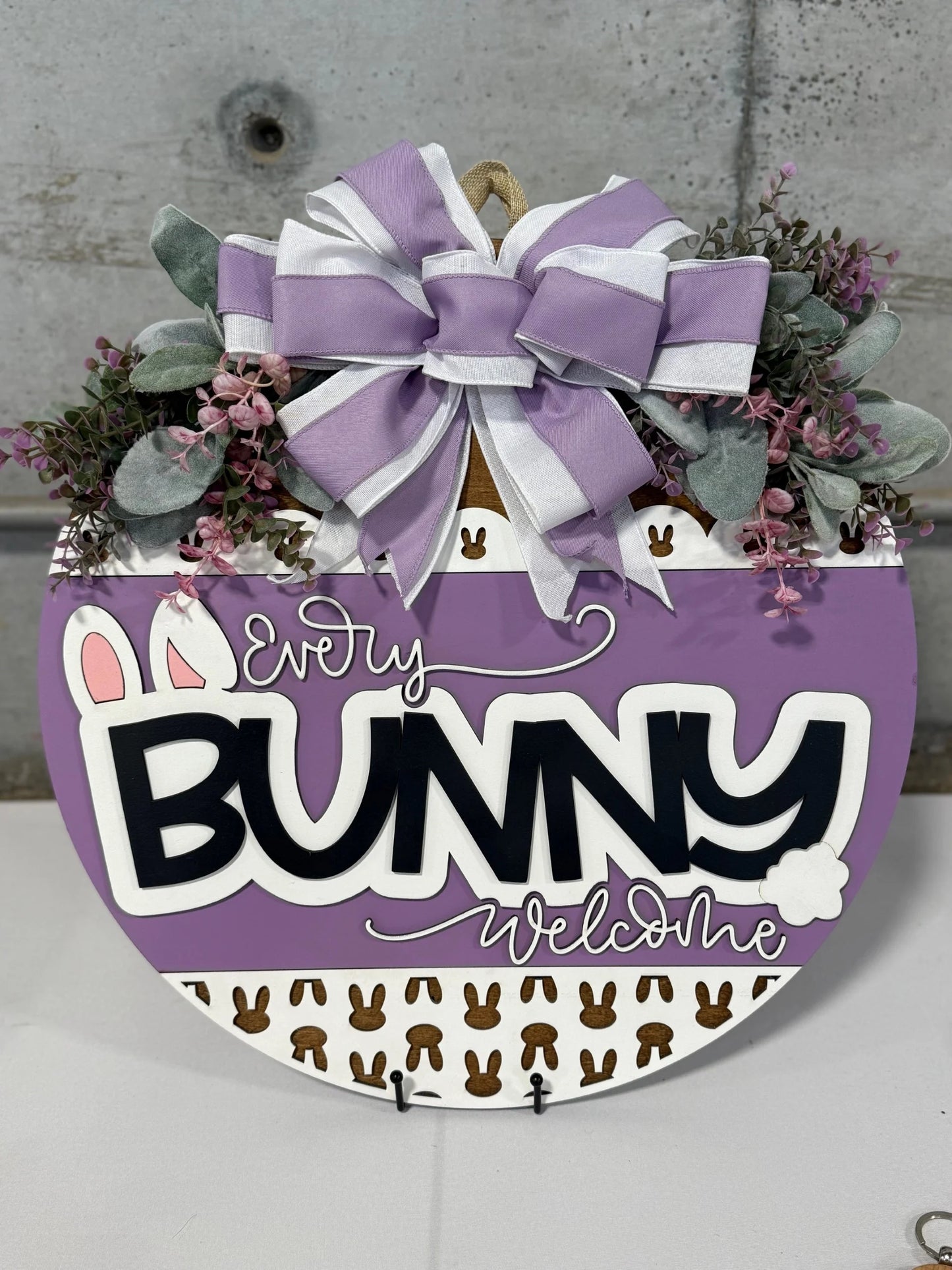 A round door hanger with the text Every Bunny Welcome in black and white on a purple background. It features bunny ears and a tail, small brown bunnies, and is adorned with a large purple and white striped bow, greenery, and pink flowers. The background is a gray concrete wall.