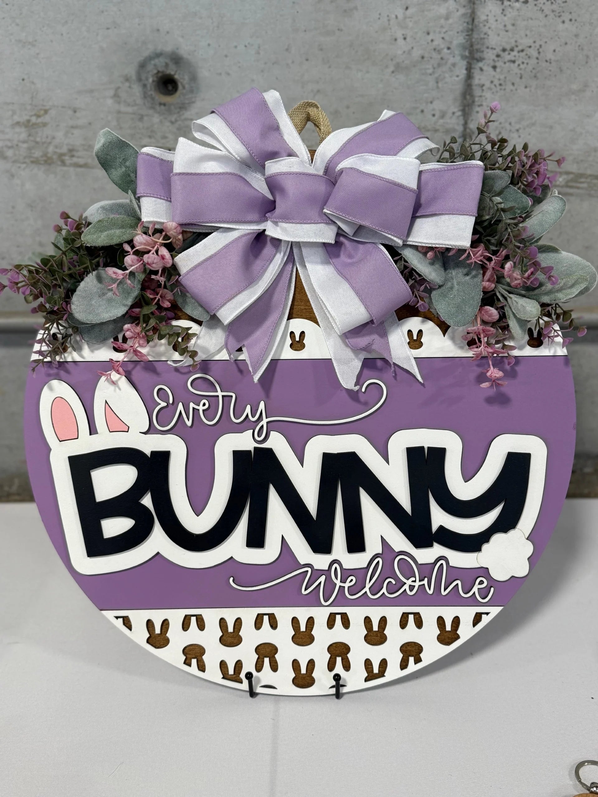 A round door hanger with the text Every Bunny Welcome in black and white on a purple background. It features bunny ears and a tail, small brown bunnies, and is adorned with a large purple and white striped bow, greenery, and pink flowers. The background is a gray concrete wall.
