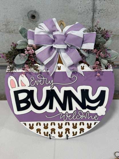 A round door hanger with the text Every Bunny Welcome in black and white on a purple background. It features bunny ears and a tail, small brown bunnies, and is adorned with a large purple and white striped bow, greenery, and pink flowers. The background is a gray concrete wall.
