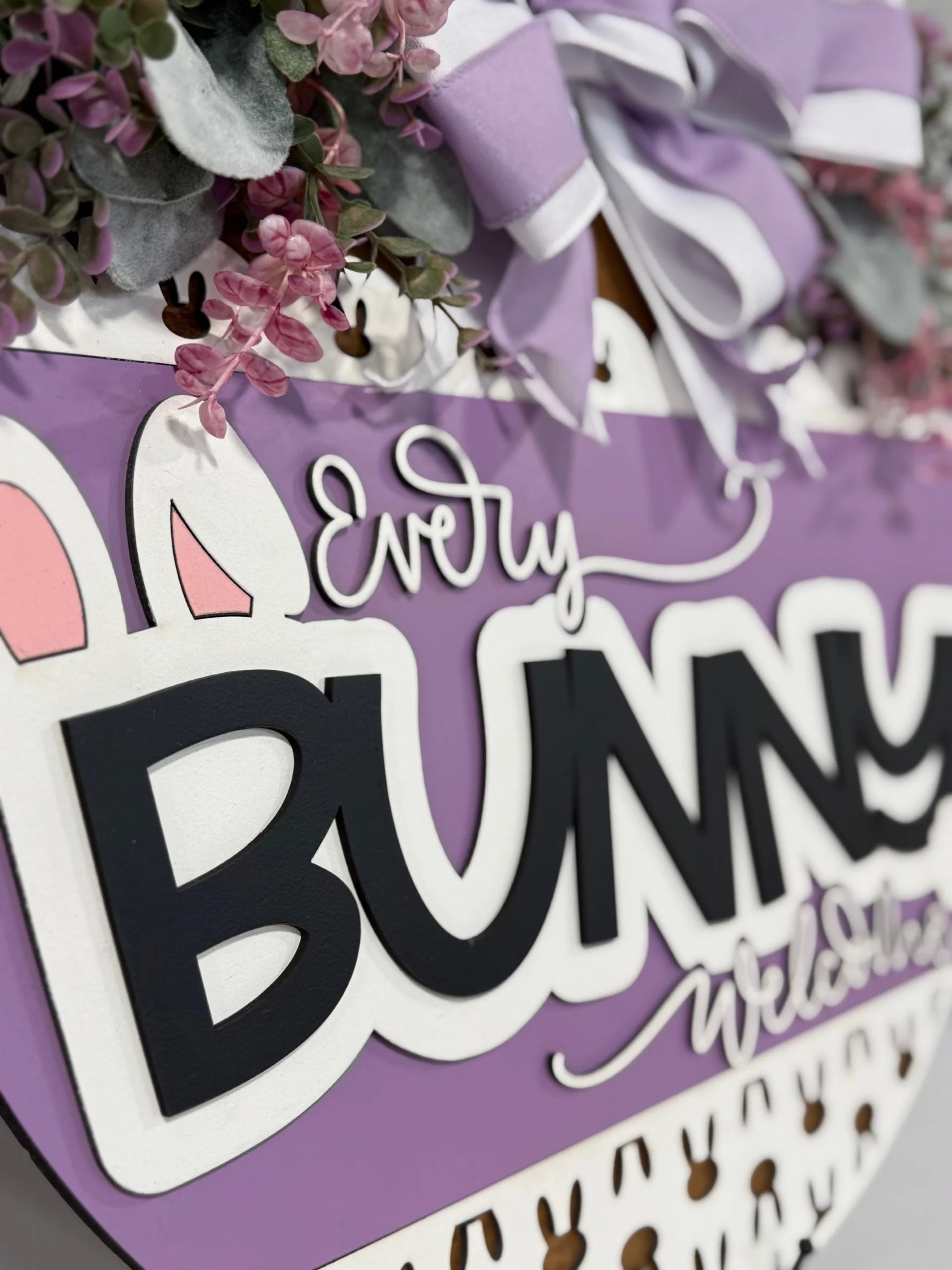 A close-up of a decorative sign featuring the text Every Bunny Welcome in playful, bold lettering. The sign is lavender with white and black text and includes bunny ear designs. Its adorned with purple ribbons and green leaves, along with small pink flowers around the top.