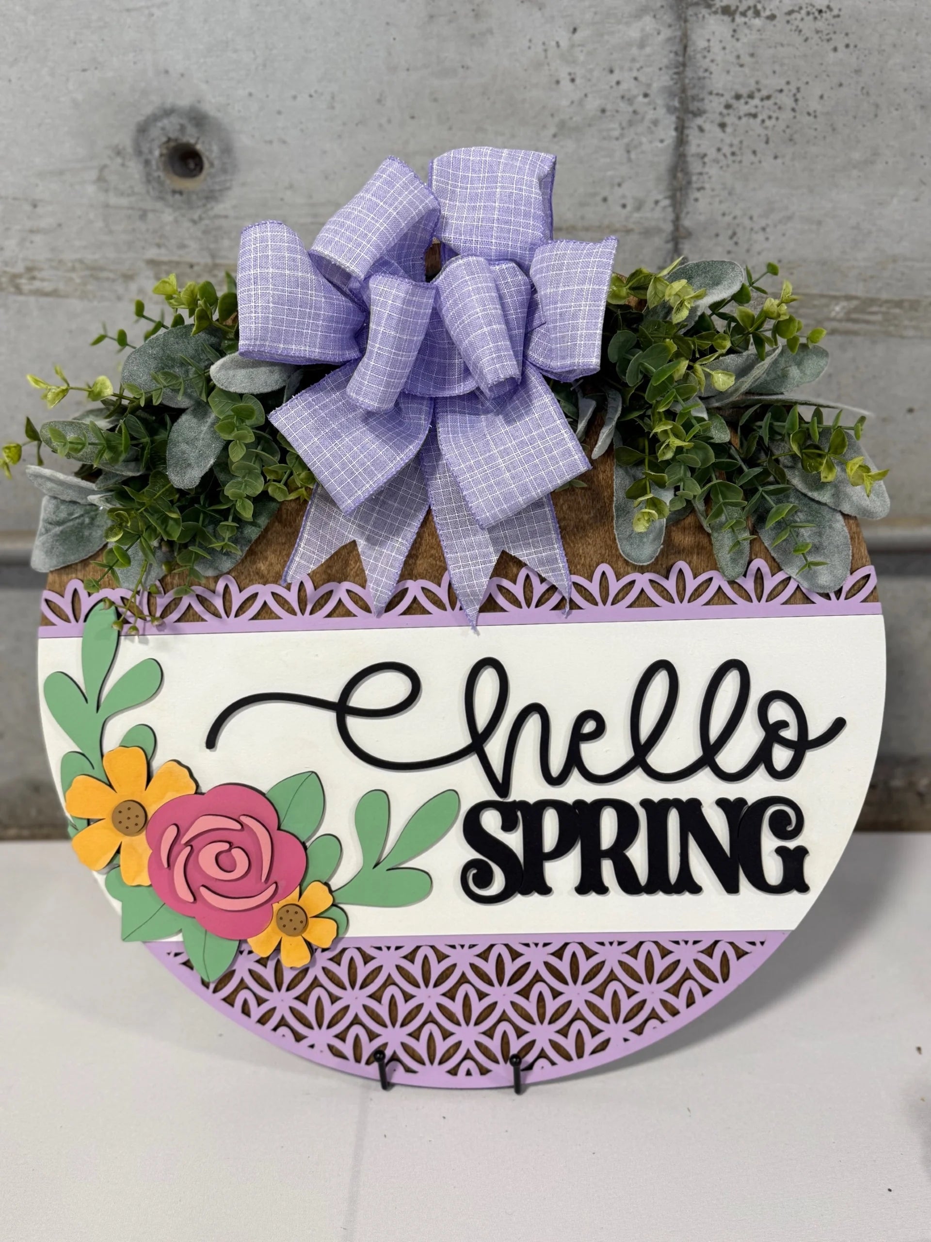 A decorative round sign with Hello Spring in black script on a white background. The lower section is lavender with a lace-like pattern. The top features green foliage and a purple checkered bow. A pink and yellow flower with green leaves are on the left side.