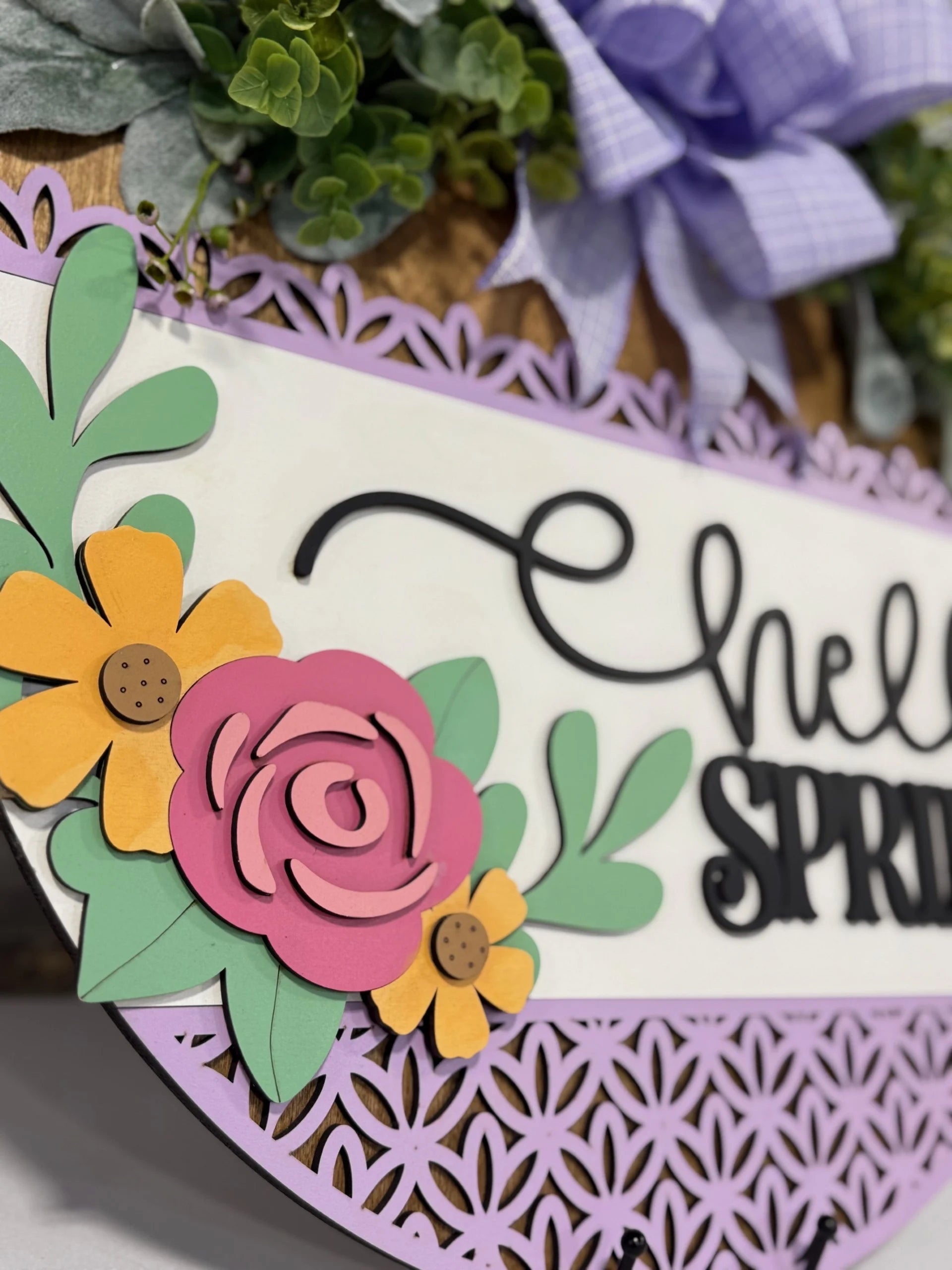 Close-up of a decorative sign reading Hello Spring with intricate black text. Surrounding the text are colorful paper flowers, including a pink rose and yellow daisies, with green leaves. The edges have a detailed lavender lace pattern. Above, theres a purple gingham bow and greenery.