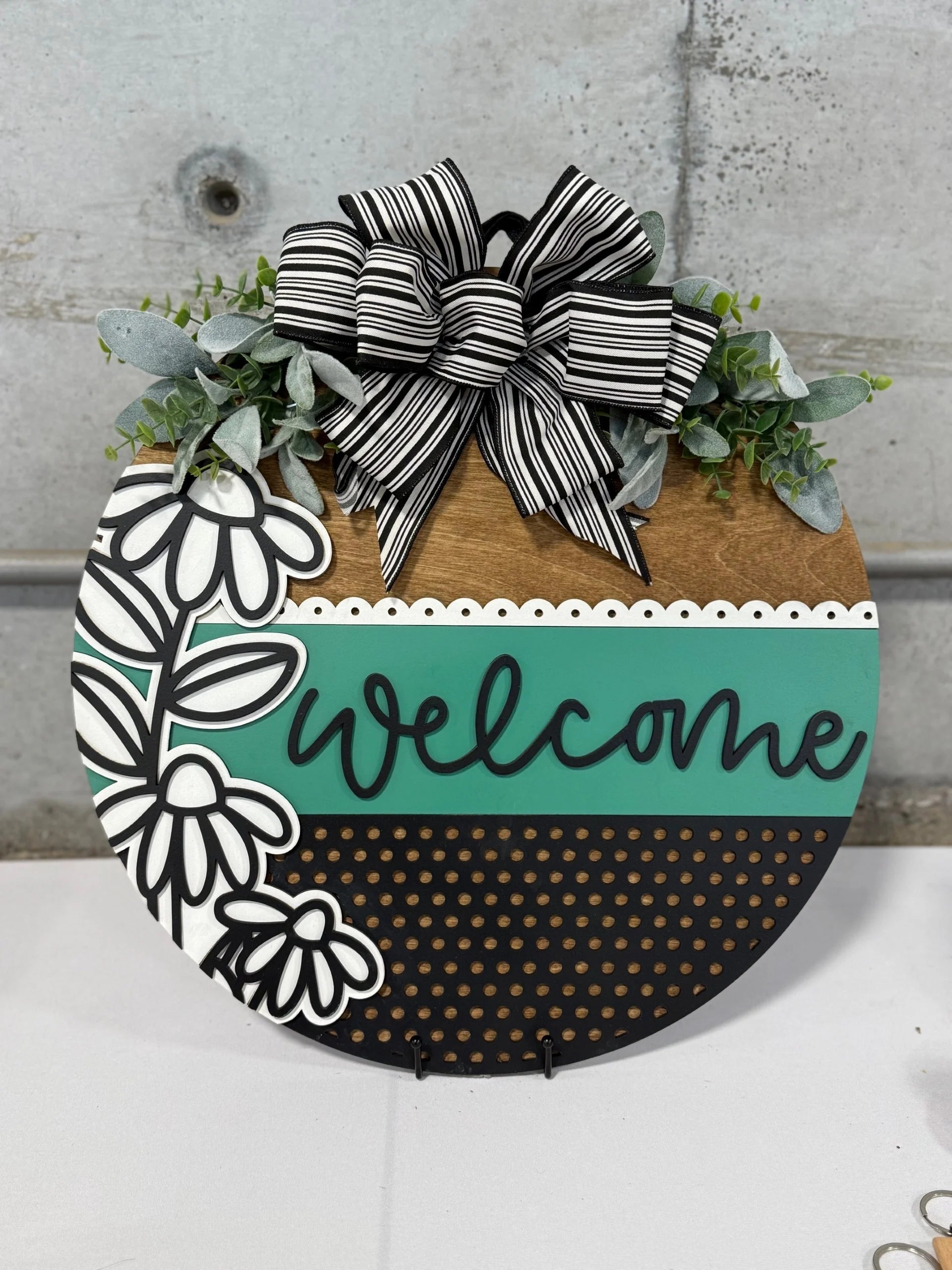 Round wooden sign with welcome text in teal on a striped, floral, and dotted design. Top section has a black and white striped bow with green foliage, including eucalyptus leaves. Background is a gray concrete wall, and the sign is displayed on a white surface.