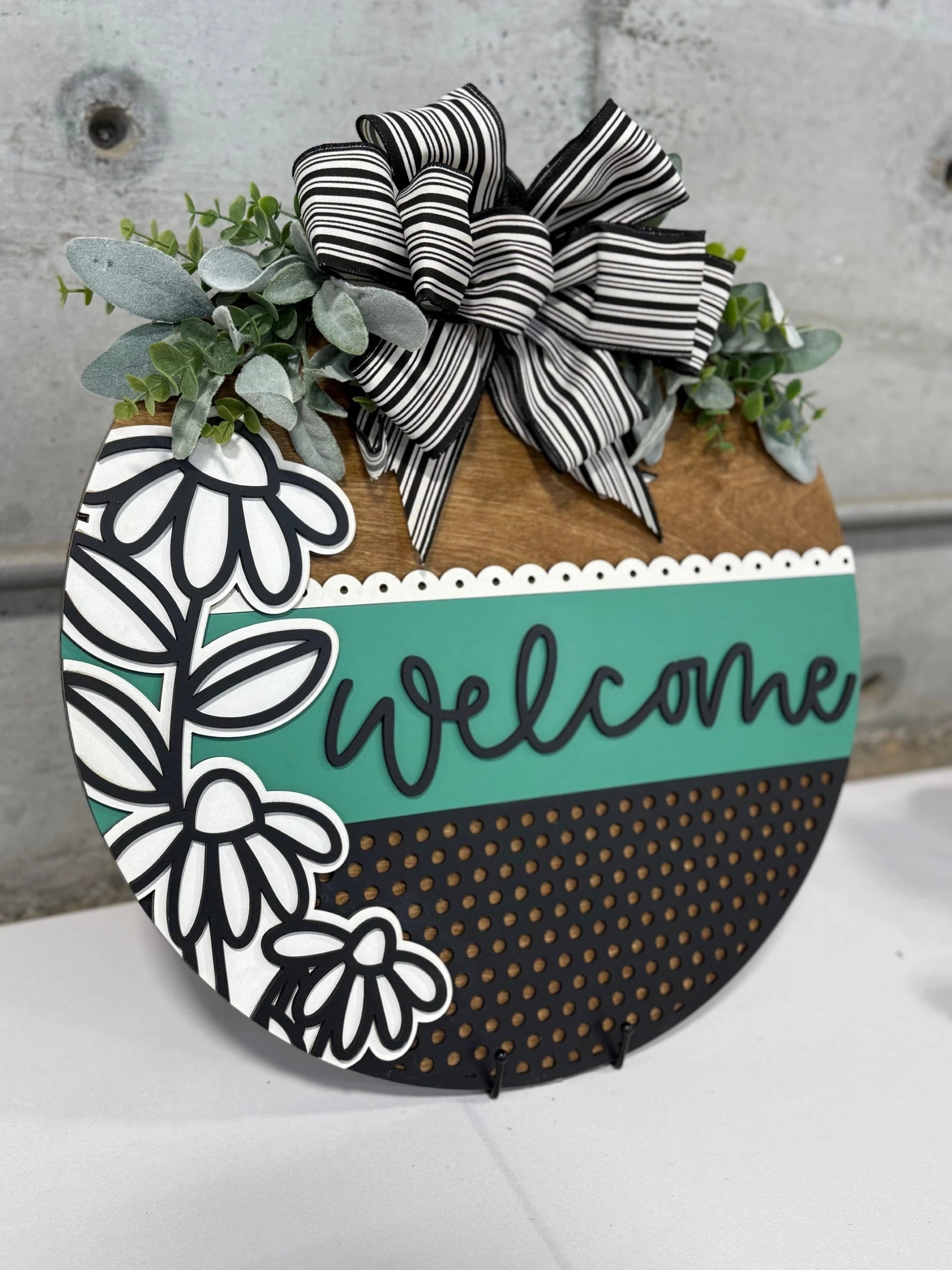 Round wooden welcome sign with a mix of designs. The top features a black and white striped bow with green foliage. Below, welcome is written on a teal stripe, next to white flowers, on a brown and black dotted background. The sign rests on a white surface.