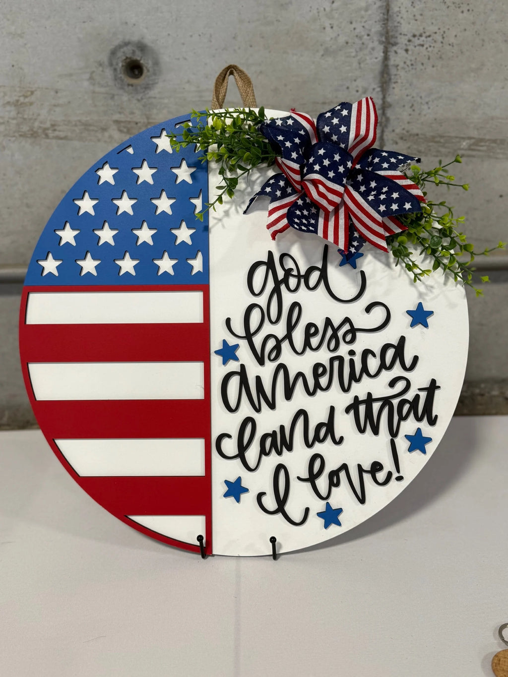 Round patriotic decoration with half an American flag and the other half white, featuring cursive text God bless America, land that I love! Adorned with a red, white, and blue ribbon and small green leaves. Displayed against a gray concrete wall.