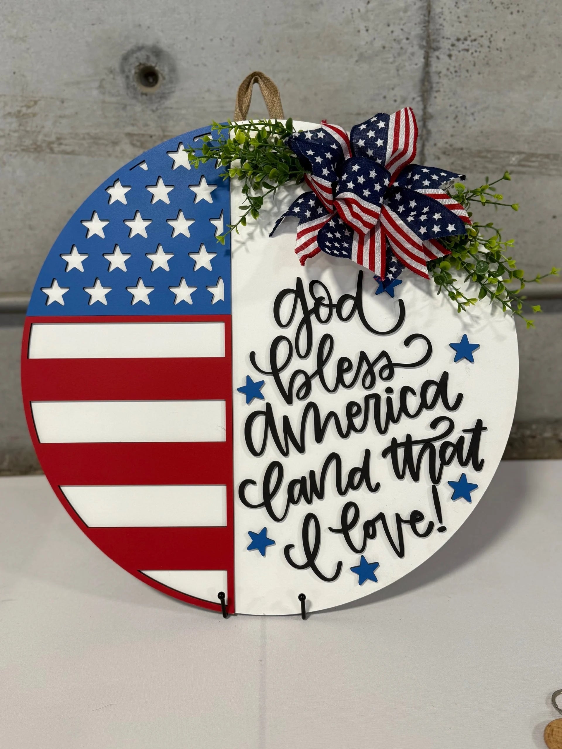 Round patriotic decoration with half an American flag and the other half white, featuring cursive text God bless America, land that I love! Adorned with a red, white, and blue ribbon and small green leaves. Displayed against a gray concrete wall.