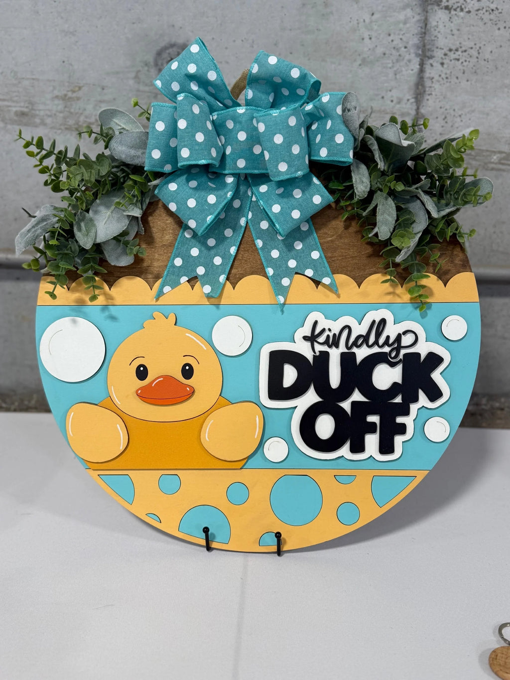 A round sign with a playful design features a yellow rubber duck and the text “Kindly Duck Off” in bold letters. The sign is decorated with a blue and white polka-dotted bow and artificial greenery at the top, all against a concrete background.