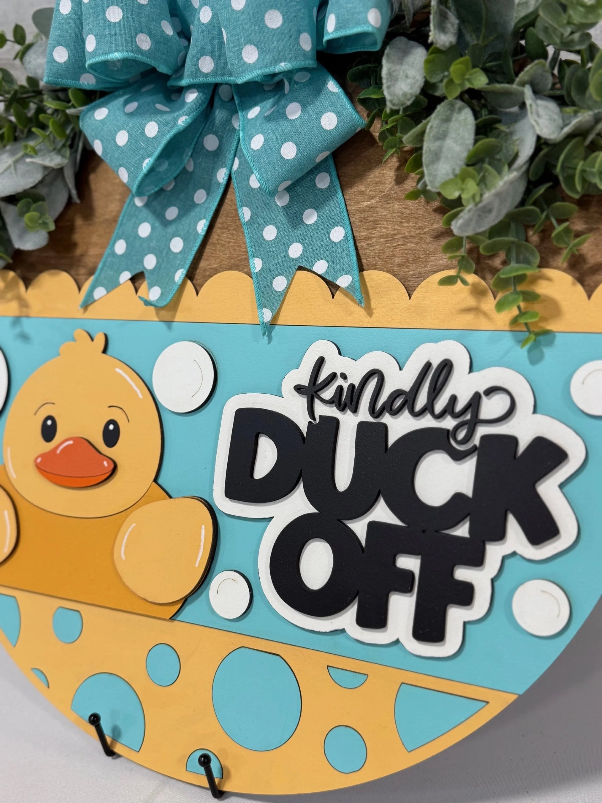Close-up of a decorative sign featuring a cartoon yellow duck next to bold text reading Kindly DUCK OFF. The sign is adorned with a large teal bow with white polka dots and bordered with green foliage. The background is a mix of aqua blue and orange with polka dots.