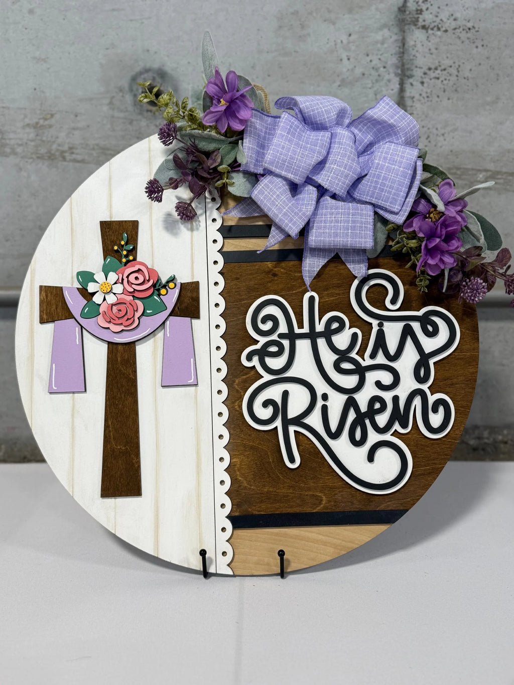 Round decorative sign with He is Risen text. Features a wooden cross adorned with pink and yellow flowers, purple fabric drapes, and a lilac bow. Purple flowers and greenery embellish the top. Background has wood and white paneling. Sign rests on a small stand.