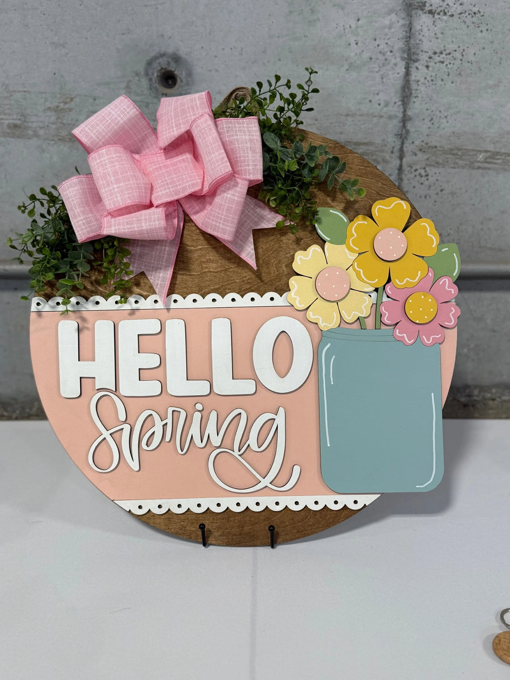 Round wooden sign with Hello Spring in white letters on a pink panel. A blue vase holds yellow and pink flowers on the right. A pink plaid bow and green foliage are on top. The background is a gray wall, and the sign is on a white surface.