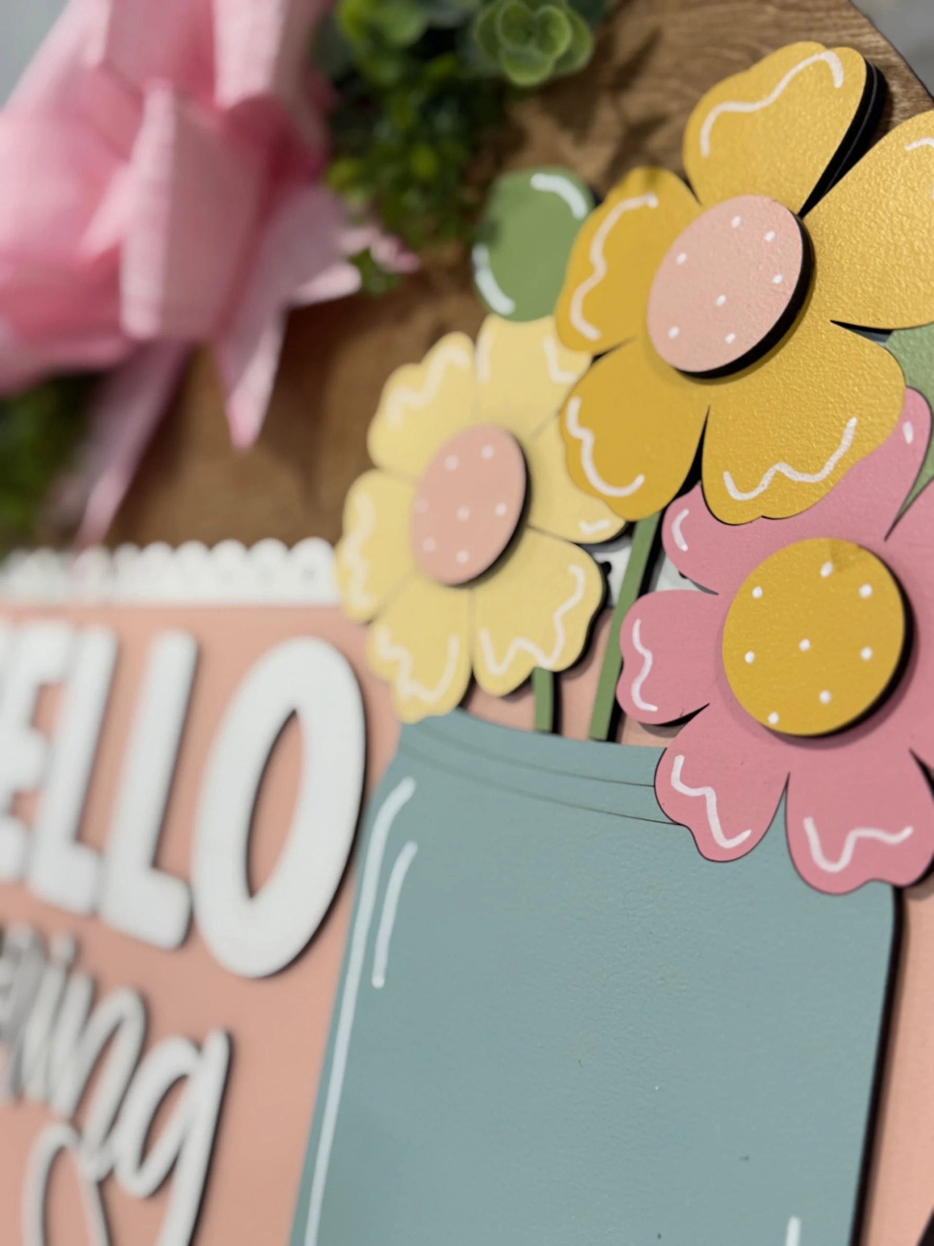 Close-up of a decorative sign featuring pastel paper flowers in pink and yellow with polka dots. The flowers are in a teal jar. To the left, the word “HELLO” is visible in white letters on a peach background. A large pink bow and greenery are in the top left corner.
