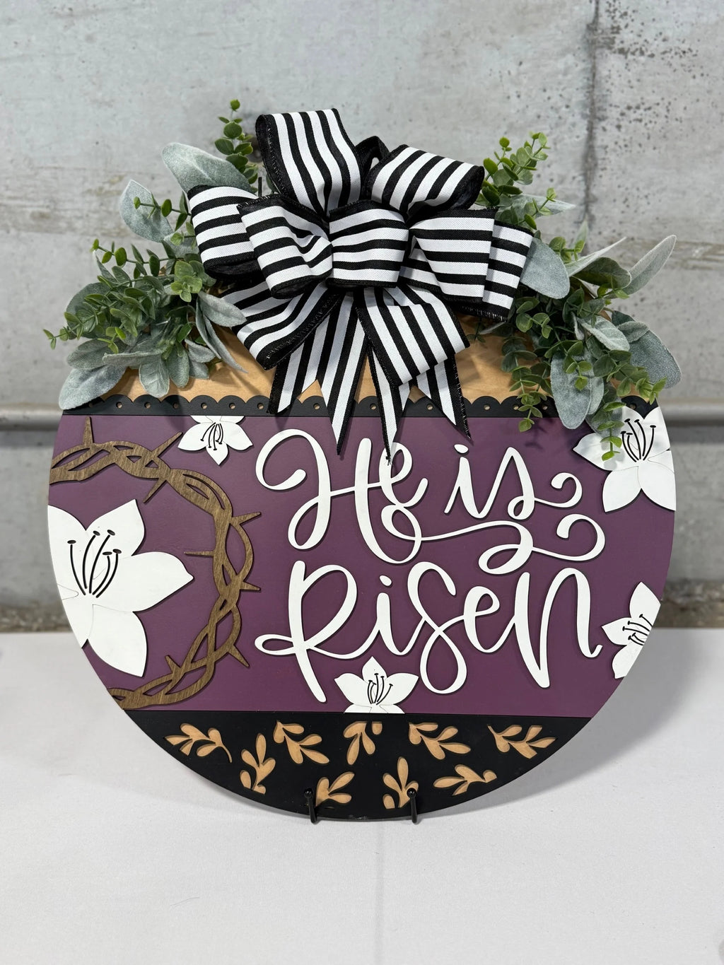 A decorative Easter sign with He is Risen written in white cursive on a purple background. It features white lilies, a crown of thorns, and a top adornment of greenery and a black-and-white striped bow. The bottom has a black section with gold leaf patterns.