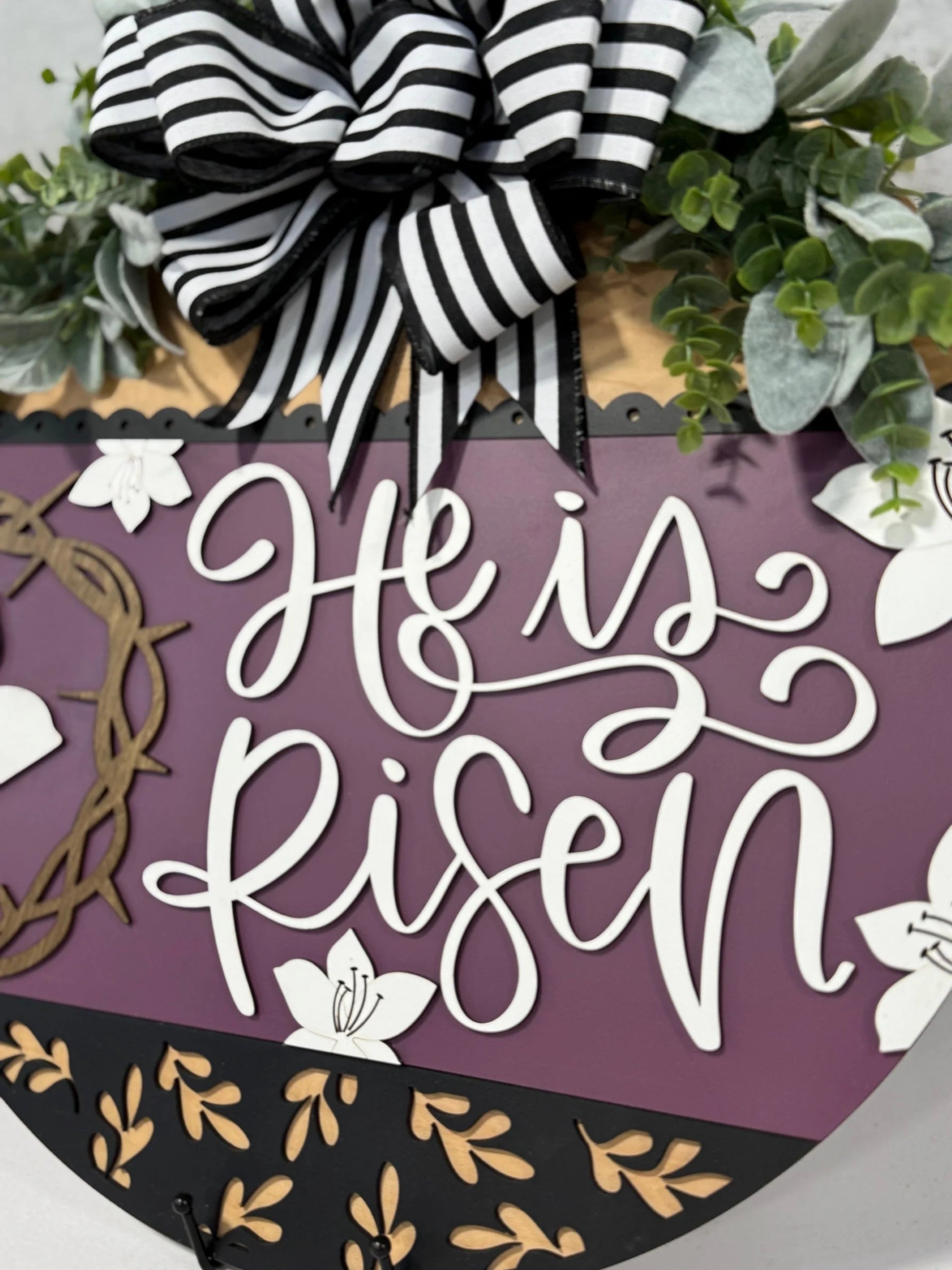 A decorative sign with the text He is Risen in white cursive on a purple background. It features a black and white striped bow at the top, surrounded by green foliage. White lilies and a brown wreath add floral accents, with a black and gold border framing the design.