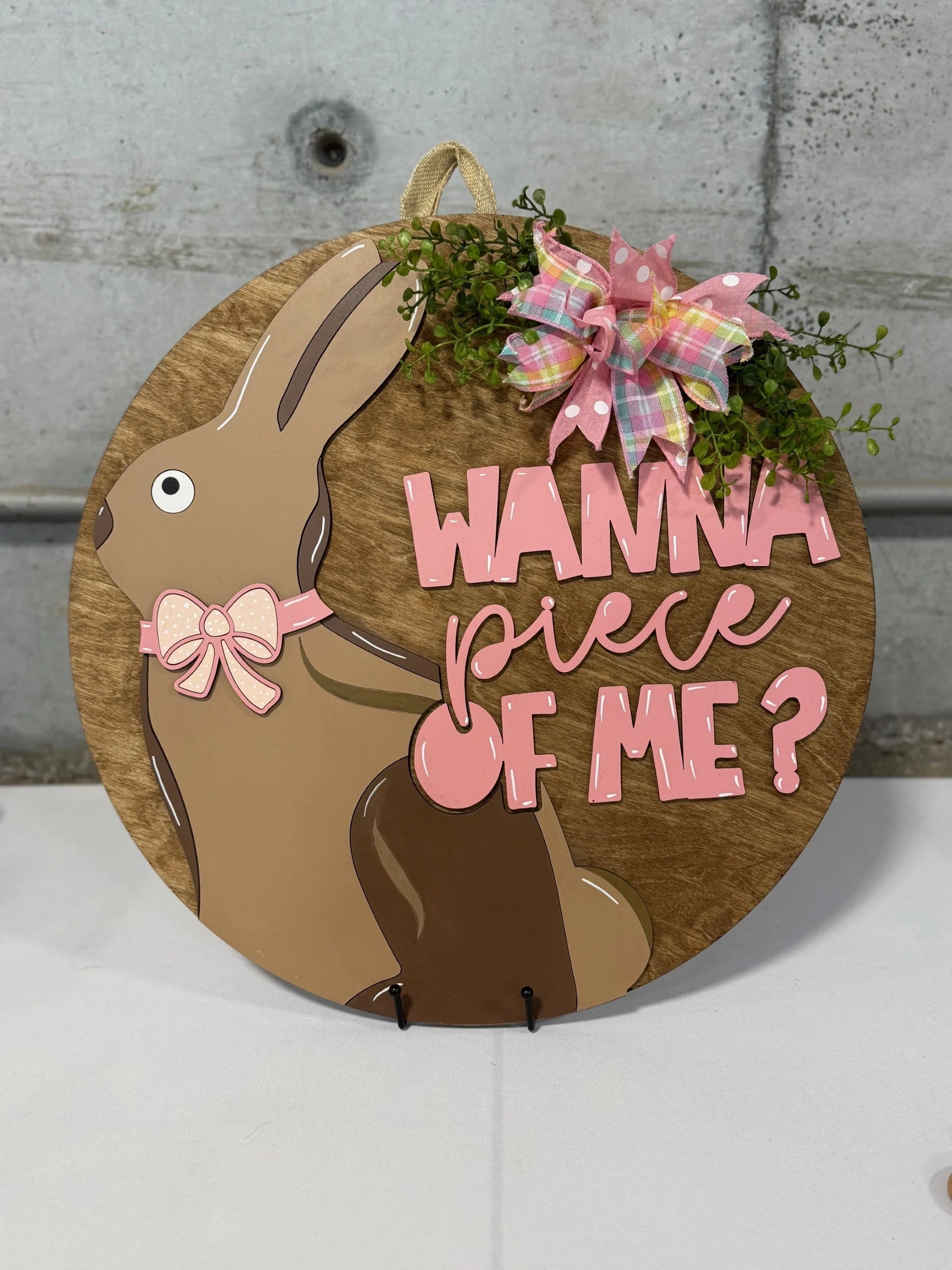 Round wooden sign with a brown bunny wearing a pink bow and a playful expression. Pink text reads Wanna piece of me? A pink and green plaid bow with greenery decorates the top right. Background is gray concrete.