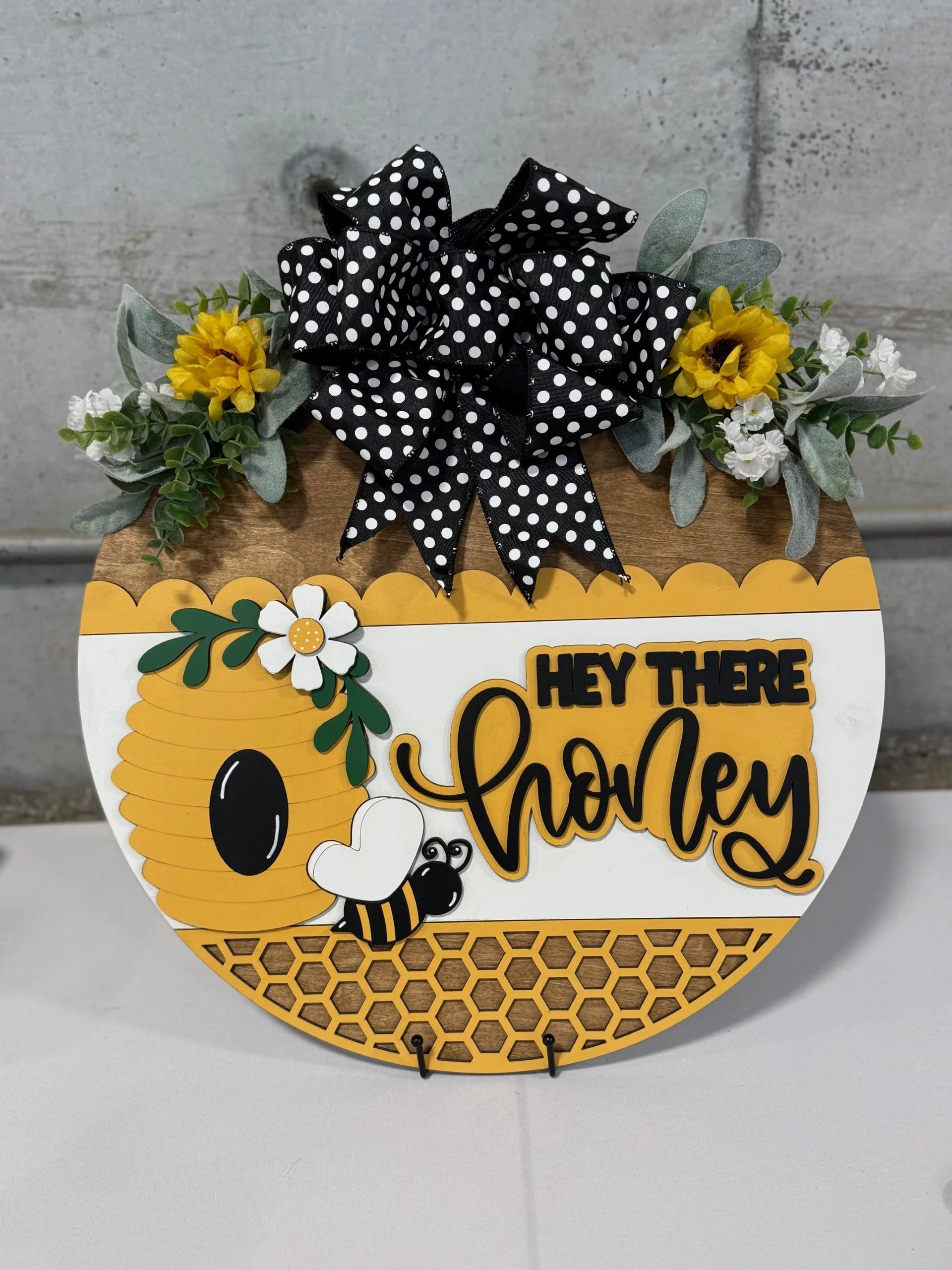 Round decorative sign with a beehive, a bee, and flowers, featuring the text Hey There Honey in yellow cursive. A black and white polka dot bow sits on top, surrounded by yellow and white flowers with green leaves. The sign has a honeycomb pattern at the bottom.