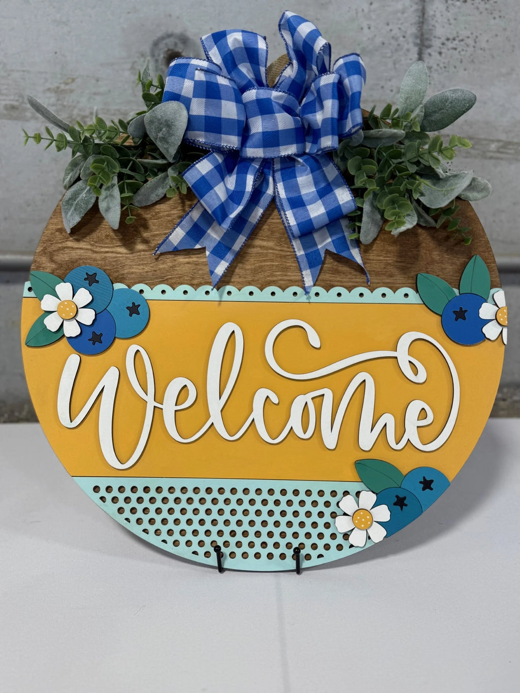 Round wooden welcome sign with a yellow stripe featuring the word Welcome in white script. Adorned with blue and white flowers, green leaves, and a blue gingham bow. Set against a gray background.