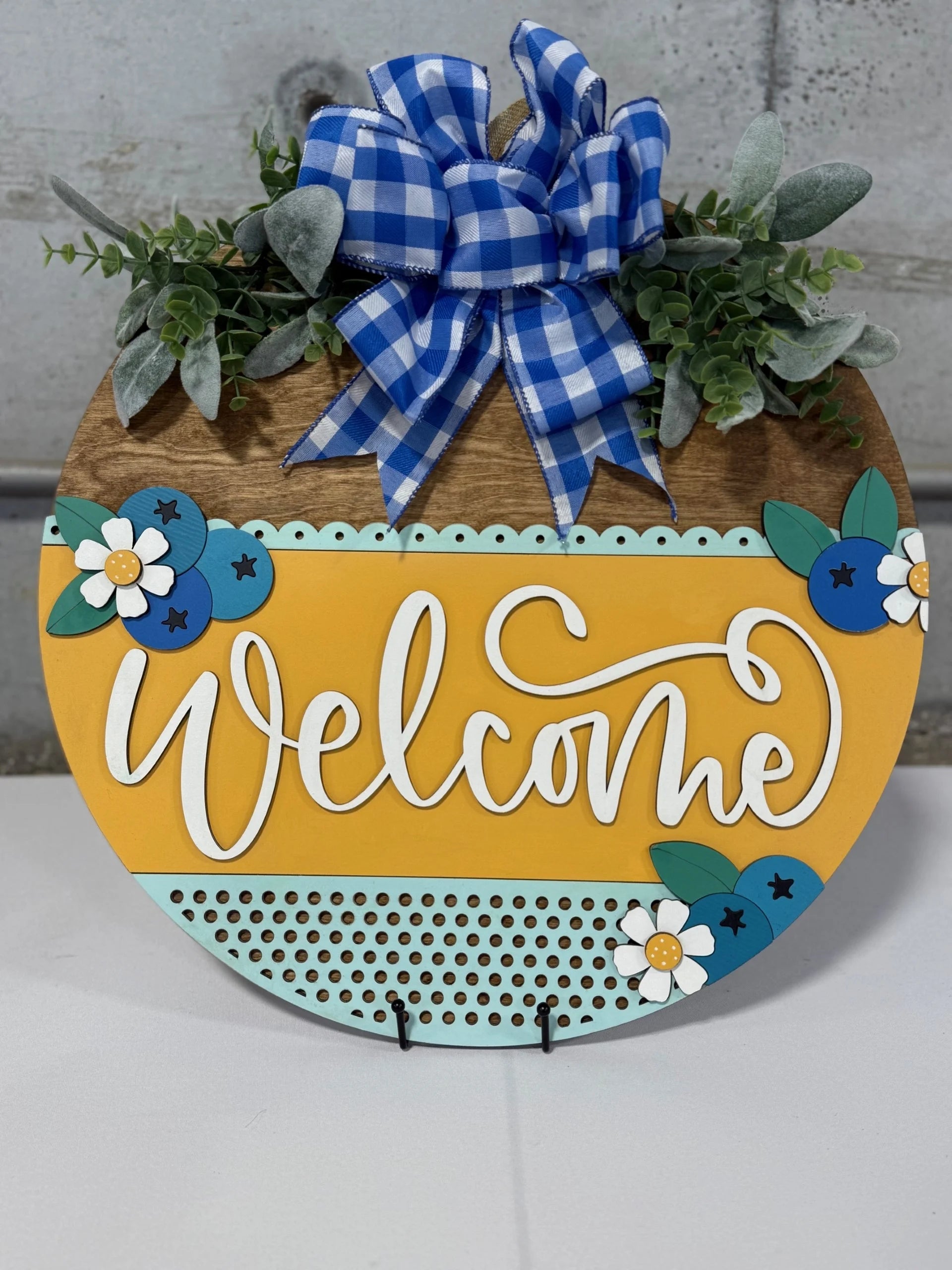 Round wooden welcome sign with a yellow stripe featuring the word Welcome in white script. Adorned with blue and white flowers, green leaves, and a blue gingham bow. Set against a gray background.