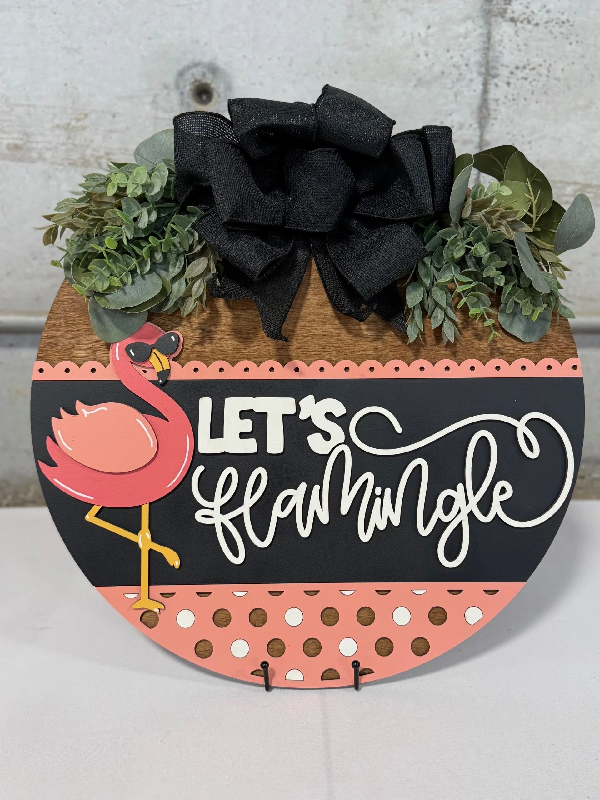 Round decorative sign with Lets Flamingle in white script. Features a pink flamingo on one leg. The background is black with a pink polka-dotted section and green leaves. At the top, theres a large black bow and more greenery. Placed on a metal stand.