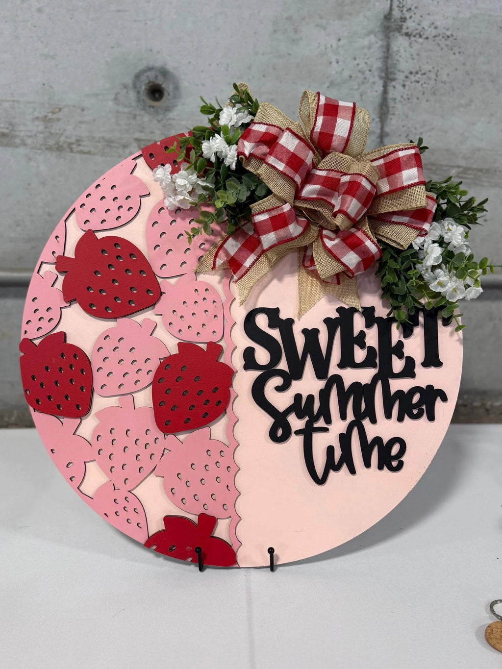 A round sign with Sweet summertime written in black cursive on a peach background. The left side features pink and red strawberry cutouts. The top right is adorned with a red and white plaid bow, burlap ribbons, and small white flowers with greenery.